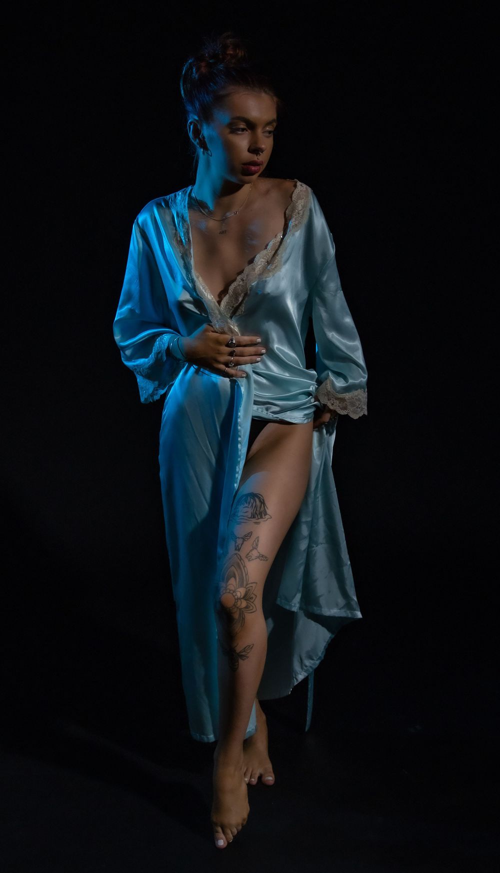 Artistic portrait in blue lighting shows a figure in a silky robe with visible tattoos against a dark background.