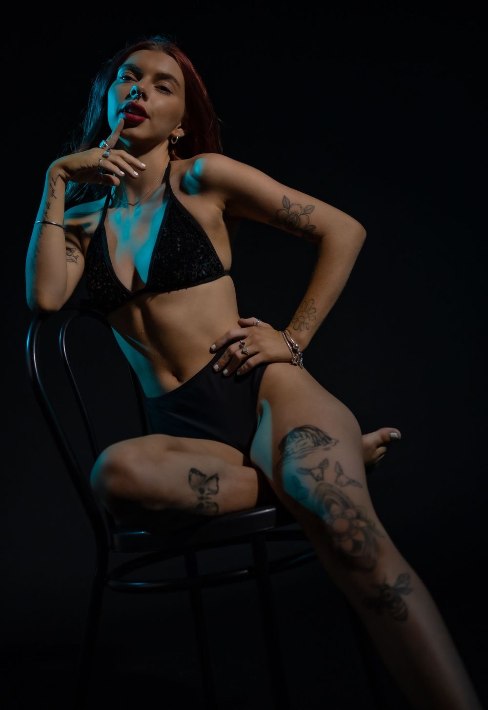 Model in black lingerie seated on chair with dramatic blue lighting and visible tattoos in dark studio setting.