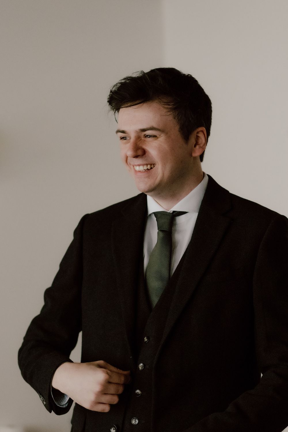 Side profile portrait in formal three piece black suit and green tie against light background.
