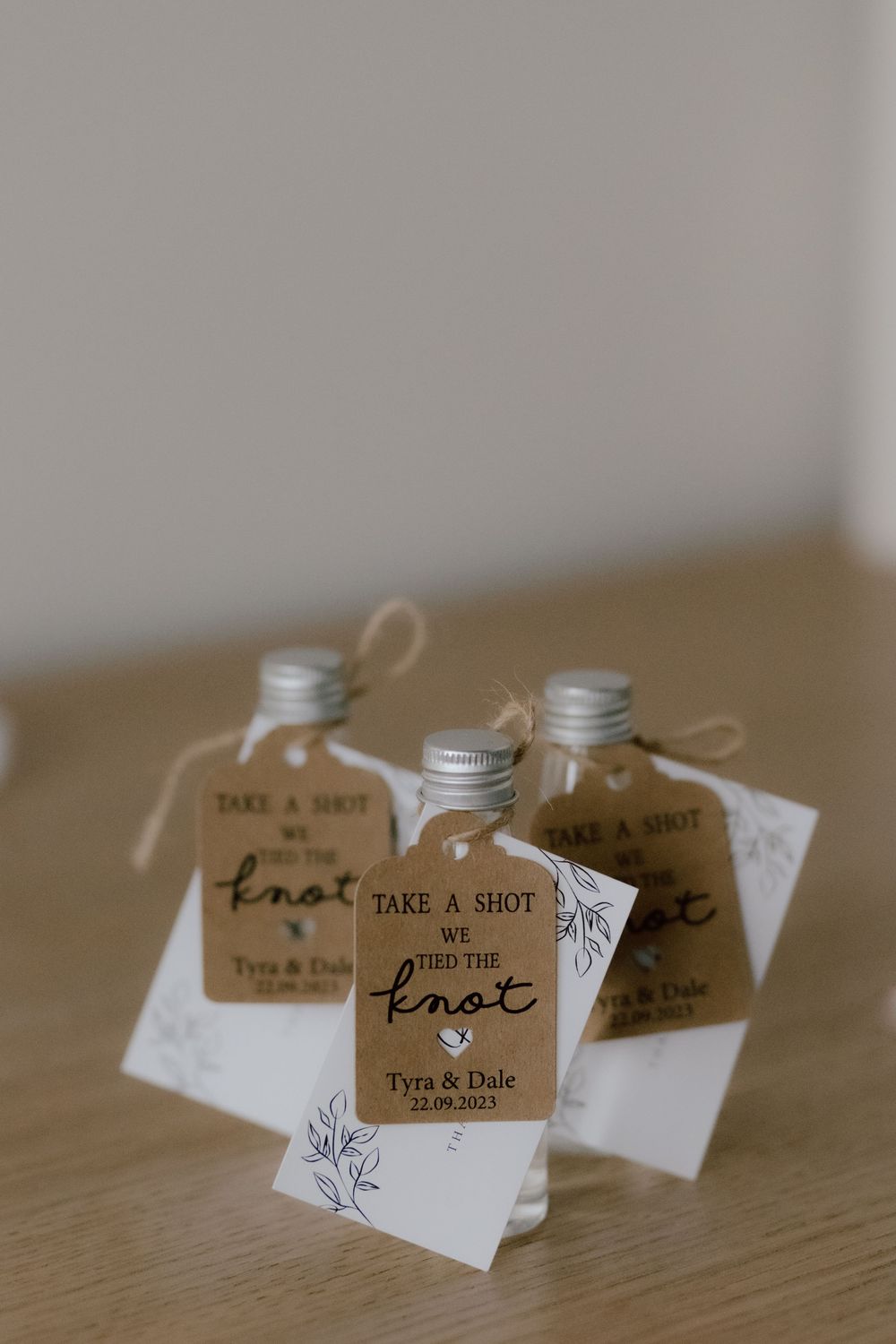 Small wedding favor glass bottles with 'Take a Shot... We tied the knot' tags and white cards on wooden surface.