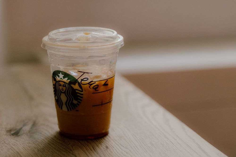 Six identical Starbucks clear plastic cups with various iced beverages sit in a row on a wooden surface.