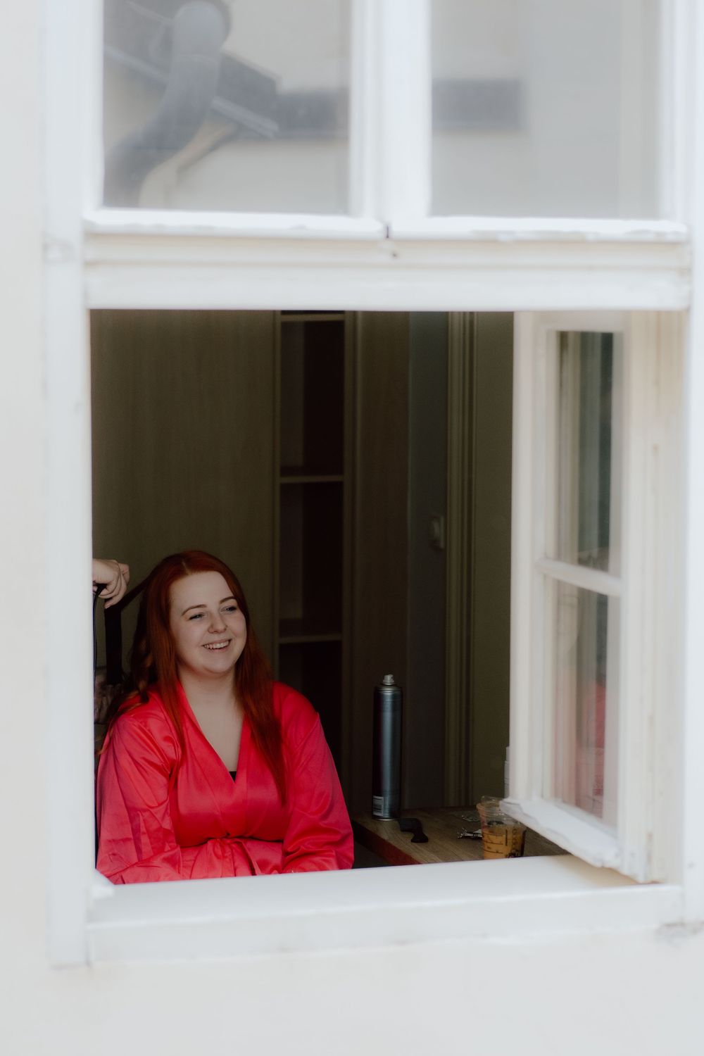 Person in vibrant red top smiles through a white window frame in a bright indoor setting.