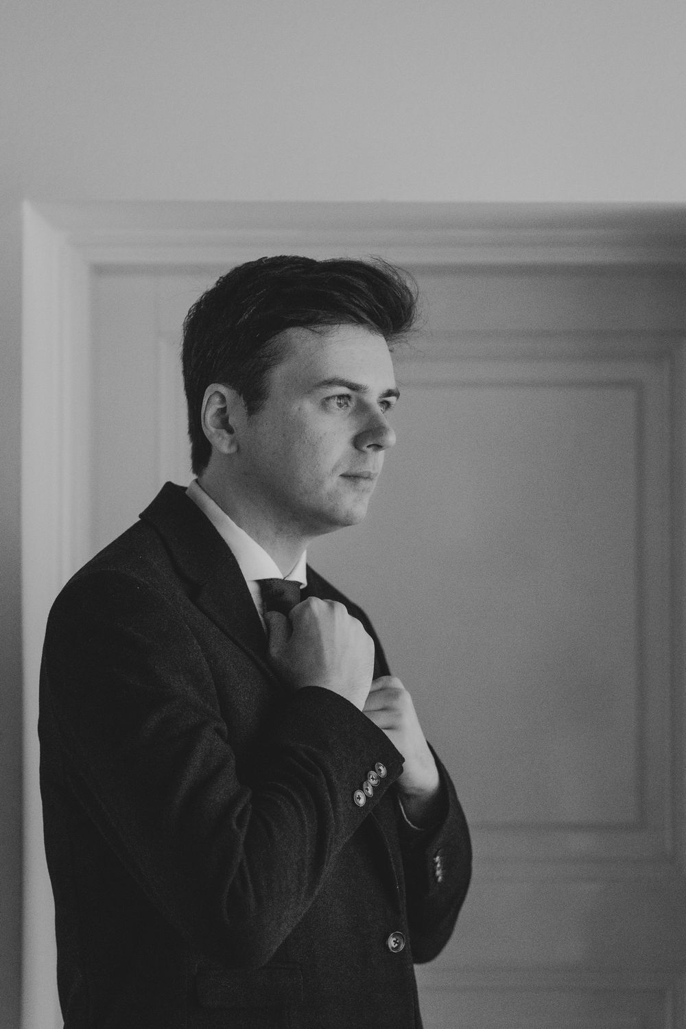 Black and white portrait of someone adjusting their tie while wearing a dark suit jacket in a formal setting.