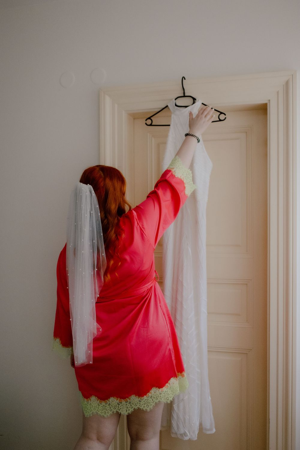 Person in red robe reaching up to hang white wedding dress and veil on door hanger in preparation for wedding day.