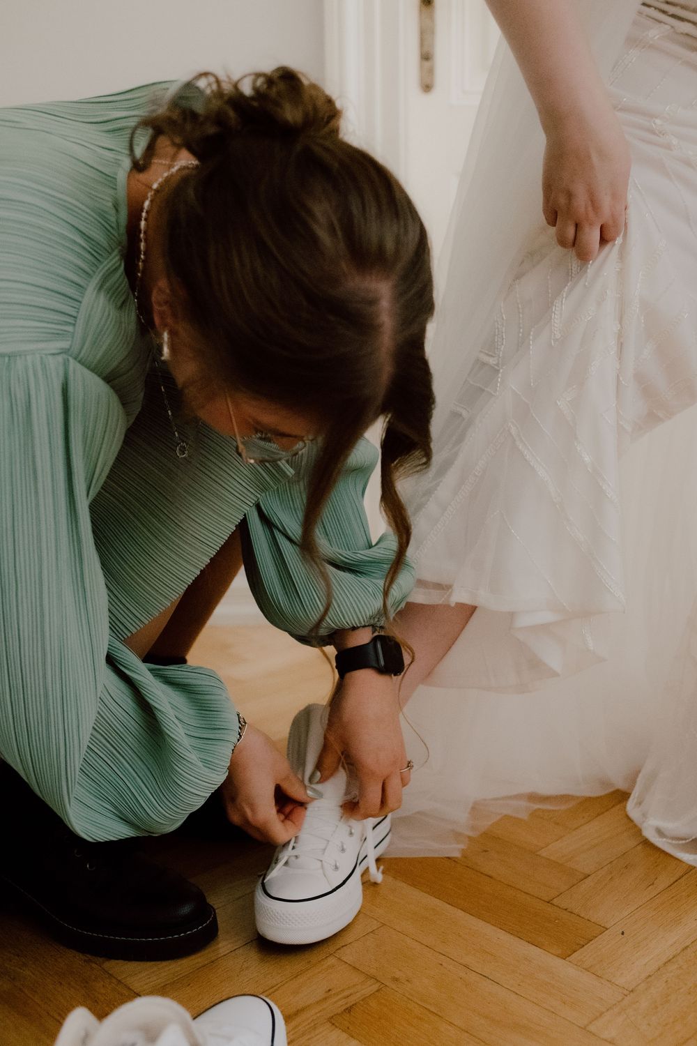 A person in a mint green dress ties white sneakers while getting ready on a hardwood floor.