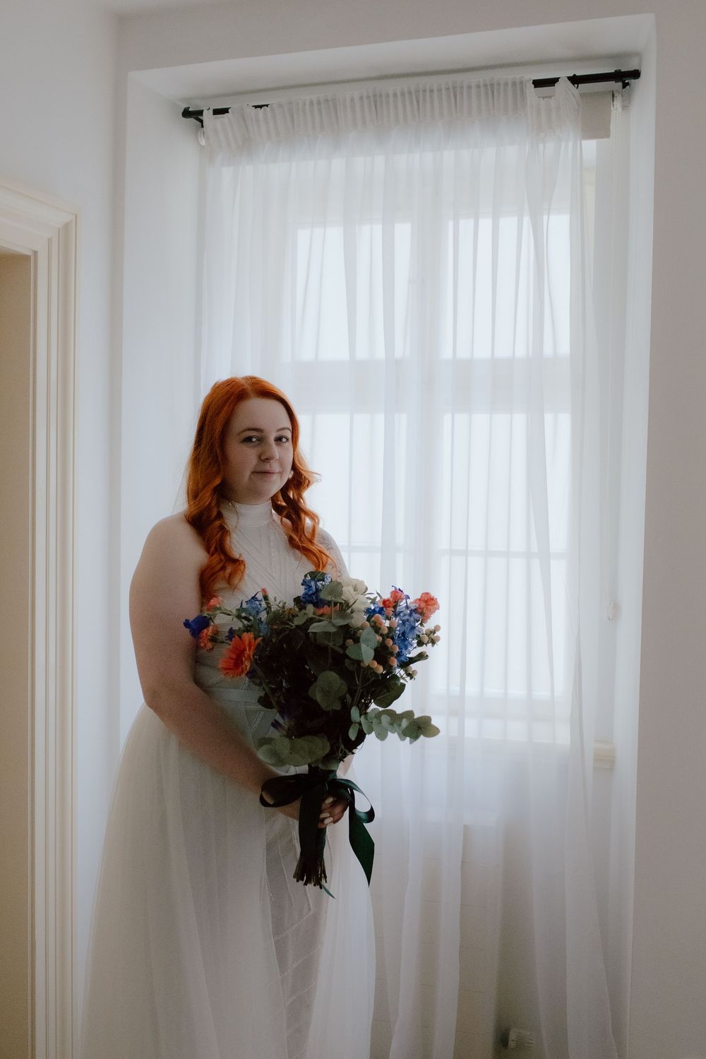 A figure in a white dress holds a colorful bouquet of flowers against sheer curtains in a naturally lit room.