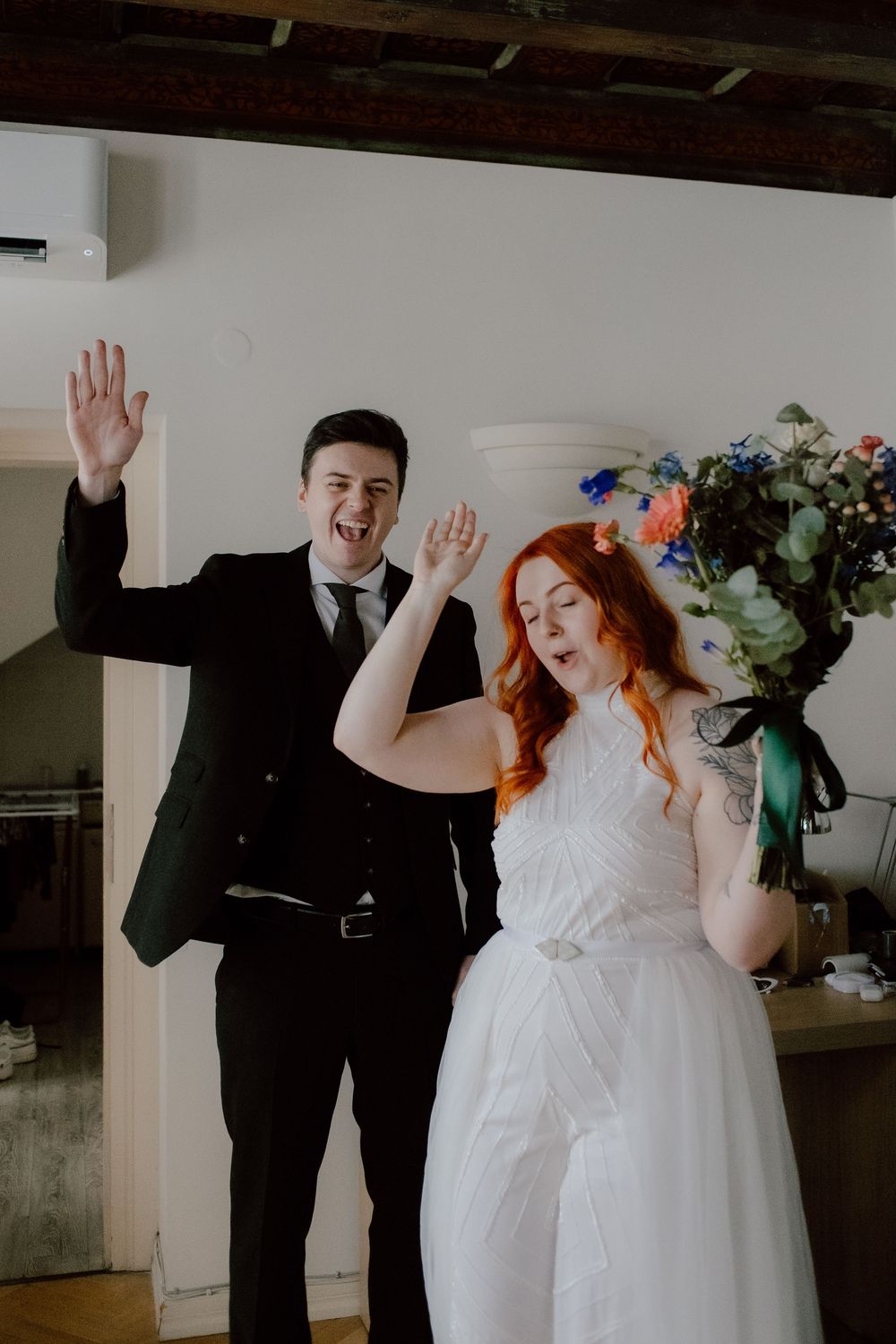A joyful newlywed couple celebrating at their wedding, with the bride in white holding a blue bouquet.