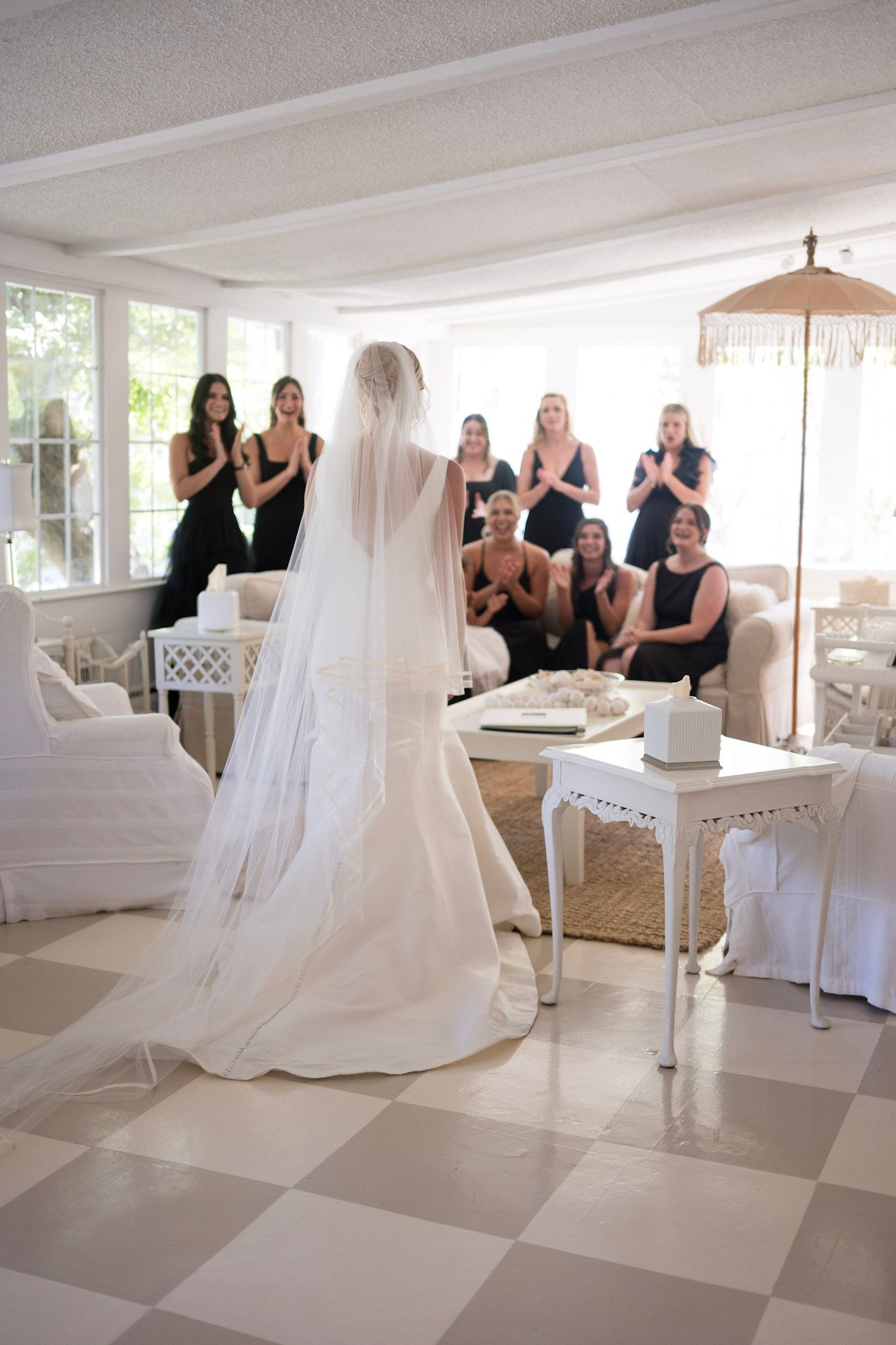 Bride in white dress revealing her gown to bridesmaids in a bright indoor setting.