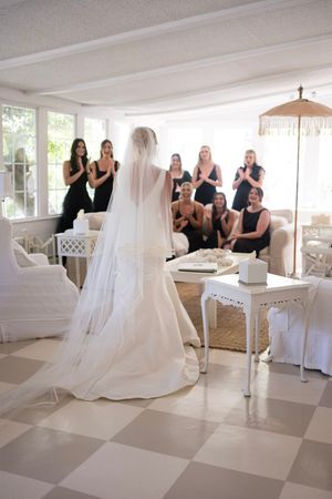 Bride in white dress revealing her gown to bridesmaids in a bright indoor setting.