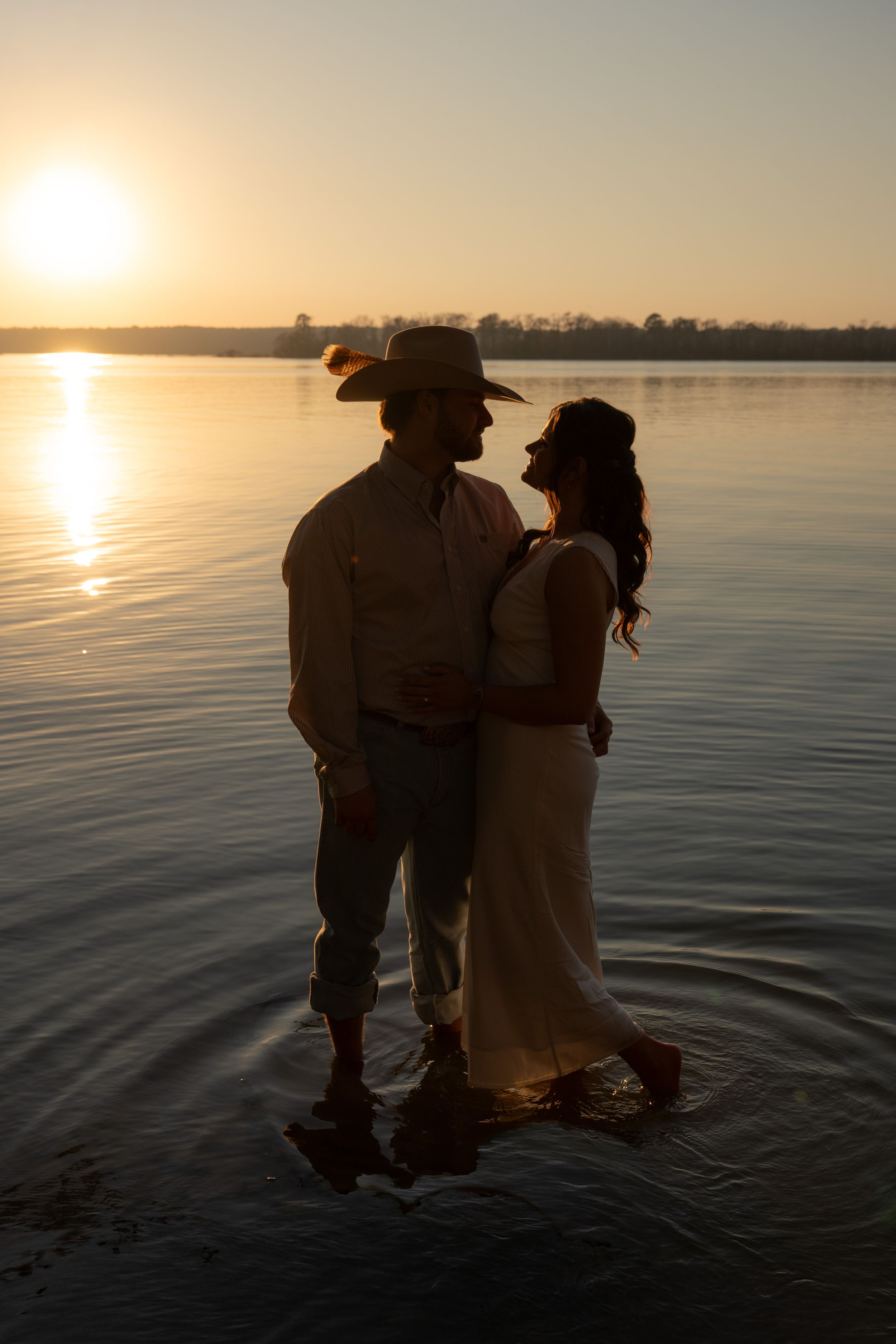 Couple standing in shallow water at golden sunset, man in cowboy hat embracing woman in white dress by a calm lake.