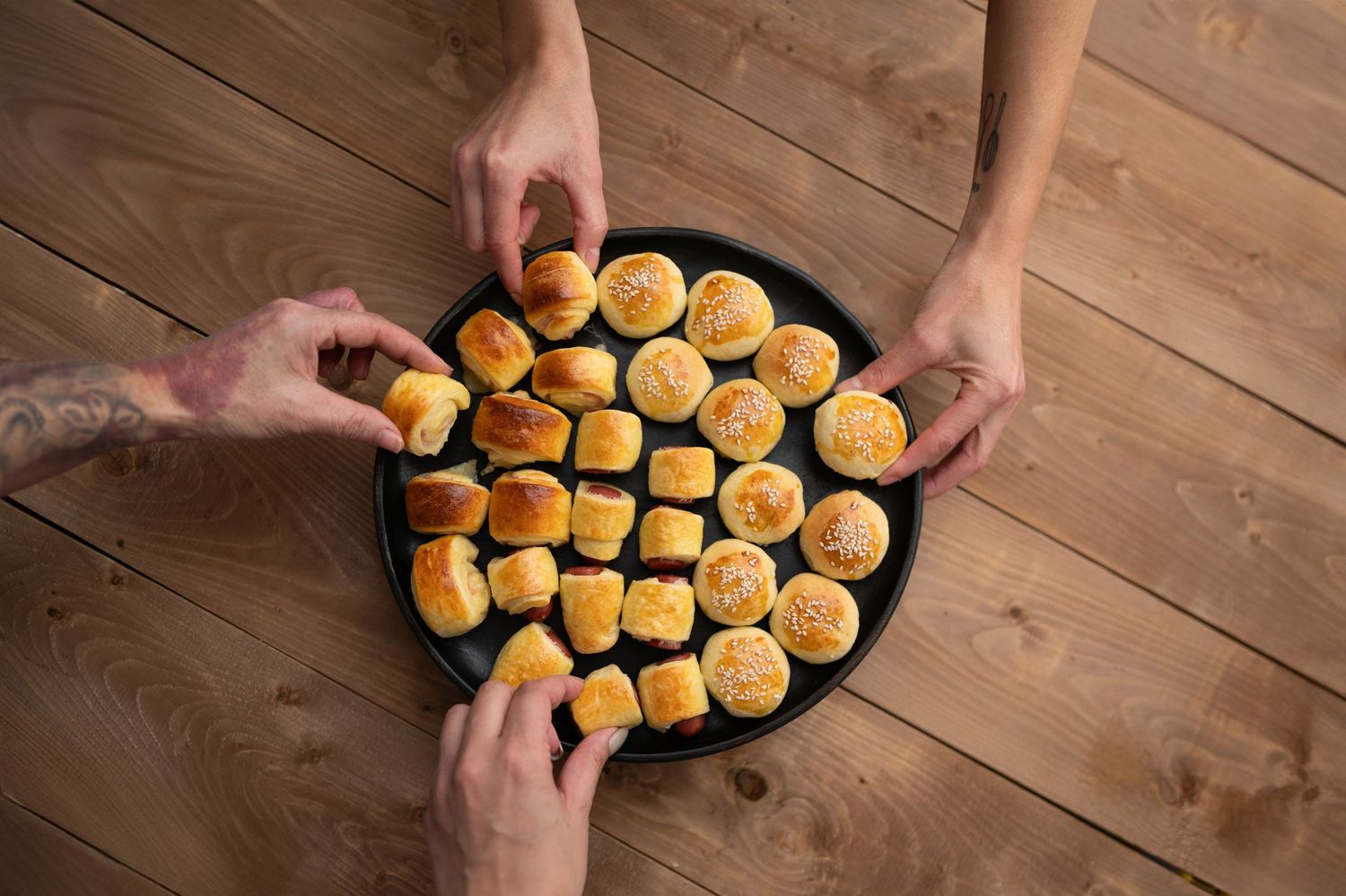 Multiple hands reaching for small golden bread rolls arranged on a dark plate.