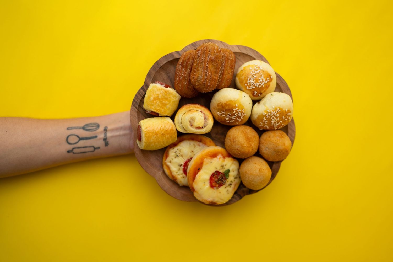 Hand holding a wooden plate of assorted bread rolls against a bright yellow background.