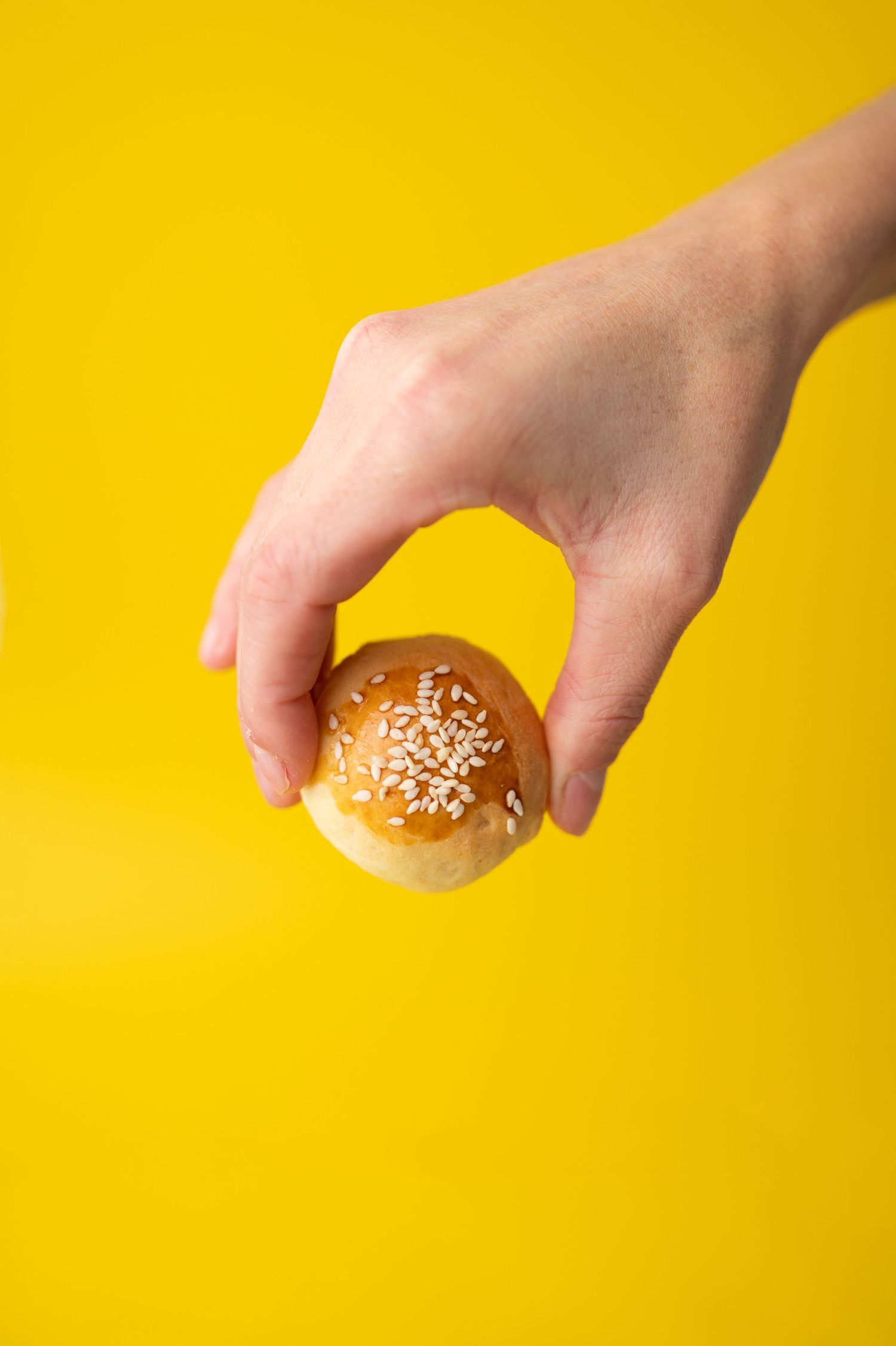 Hand holding a sesame-topped bread roll against a vibrant yellow background.