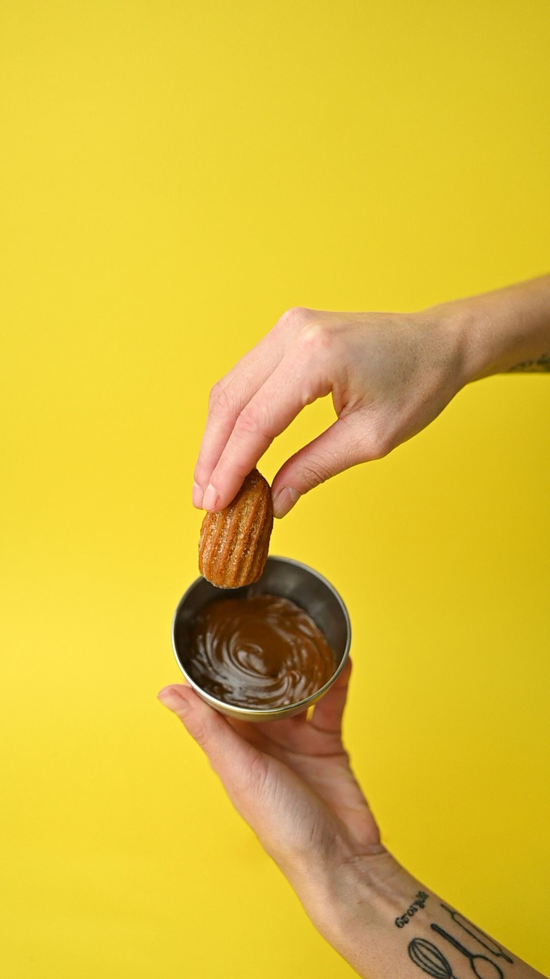 Hands dipping cookies into melted chocolate against a bright yellow background.