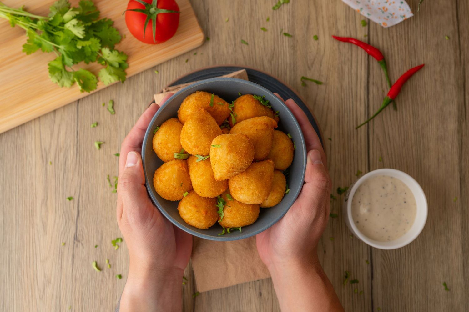 Bowl of golden breaded snacks held over wooden cutting board with fresh herbs and tomatoes.