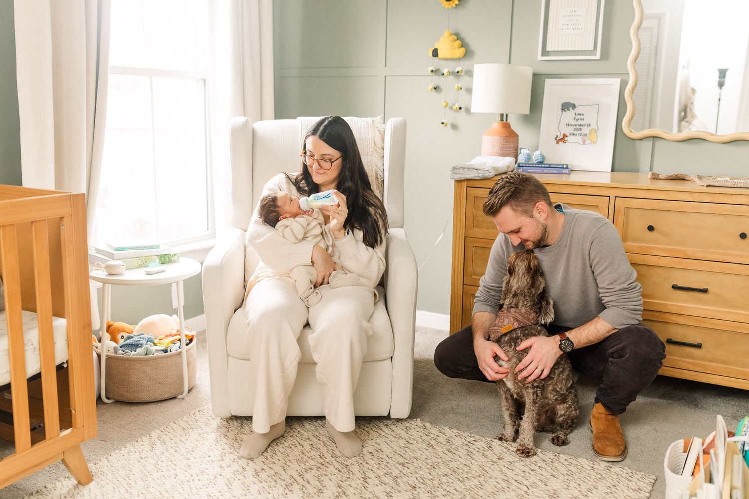 Mom feeding newborn a bottle while dad pets dog in the nursery