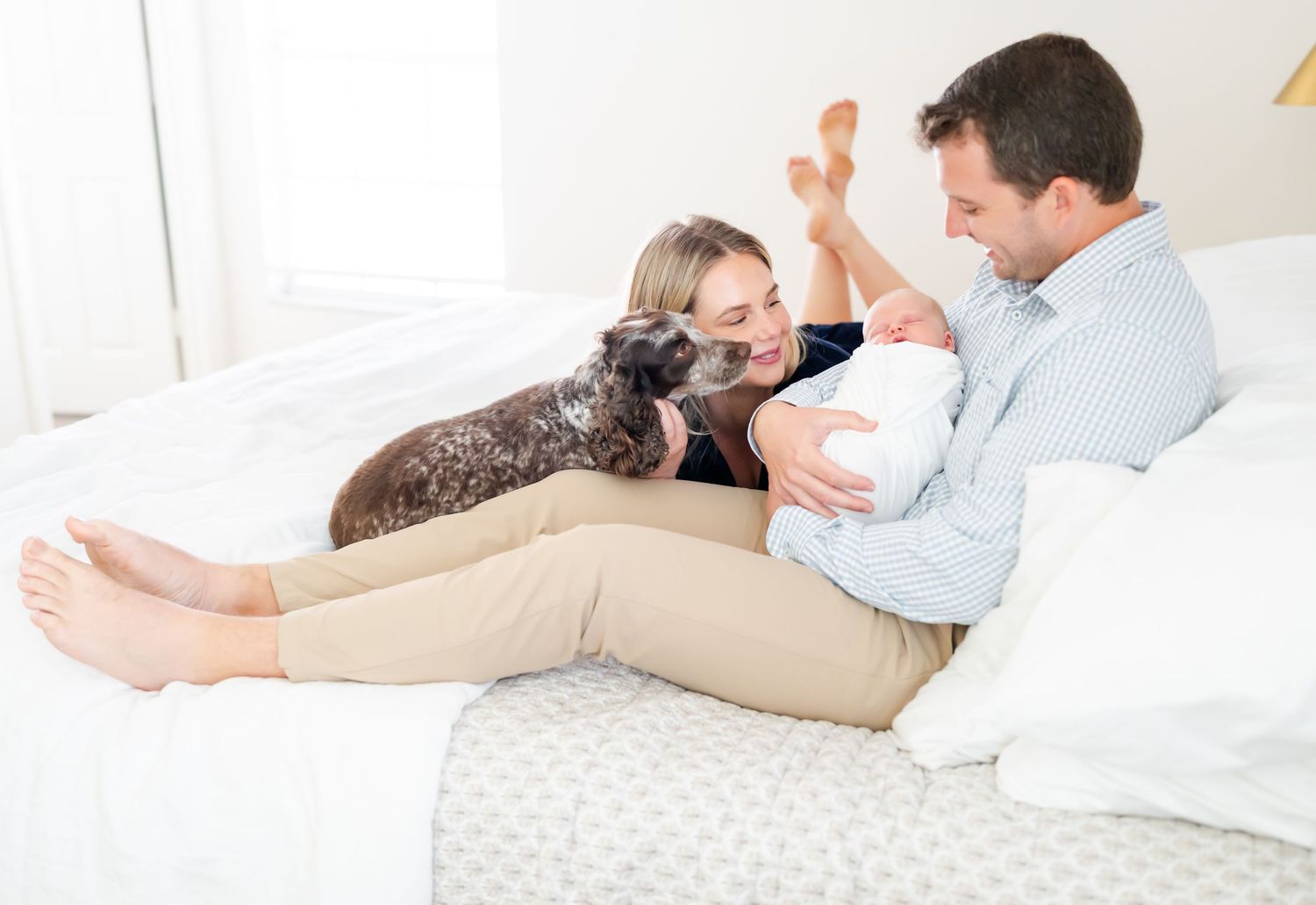 Family relaxing on white bed with dog and baby in bright, airy bedroom.