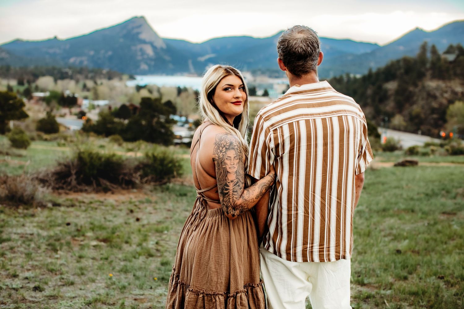 A couple in brown and striped outfits embracing in a scenic mountain landscape during a photo session.