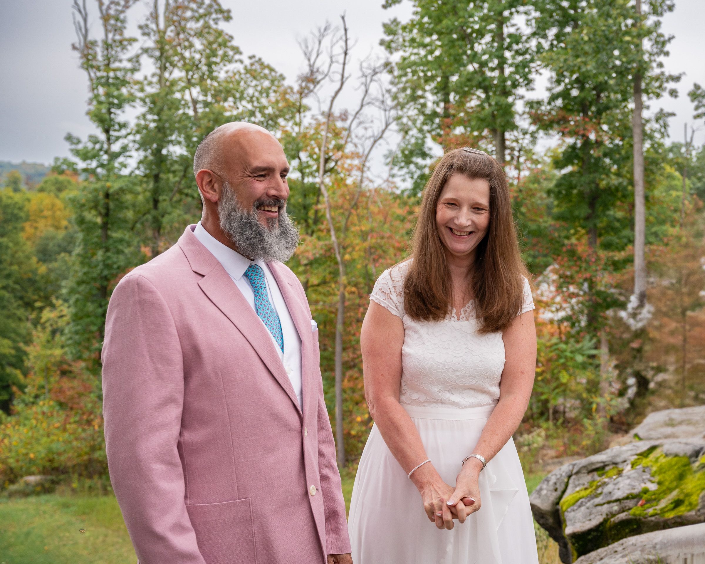 Outdoor wedding ceremony with couple in pink suit and white dress among autumn trees.
