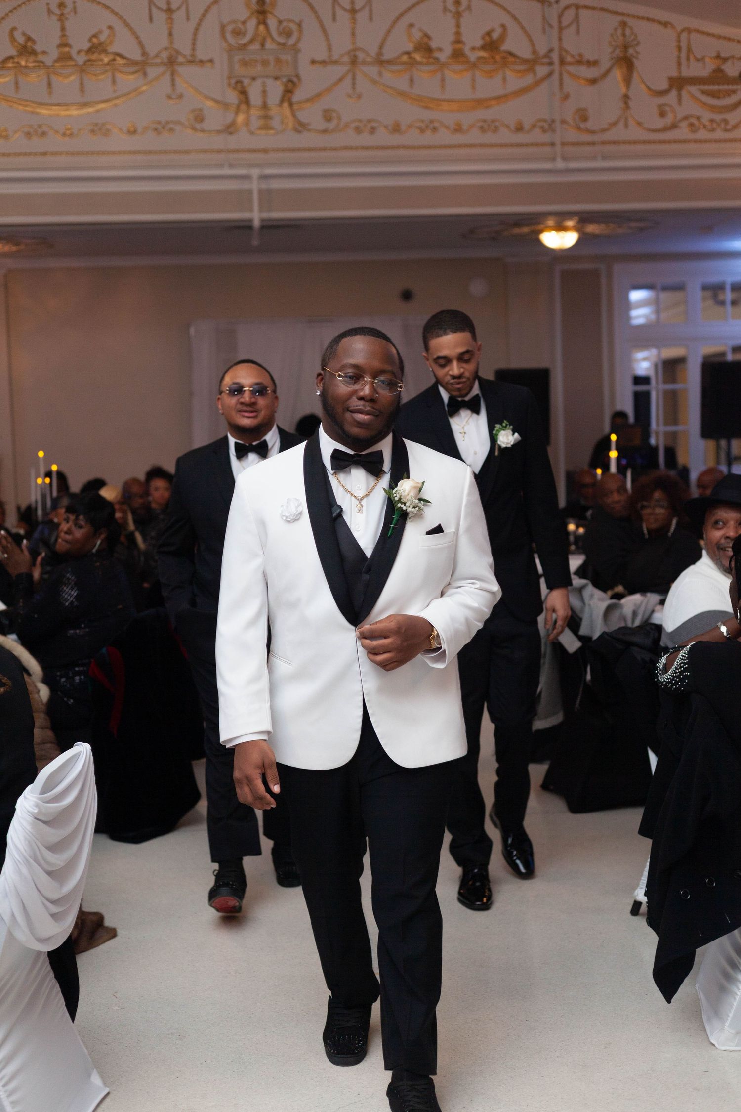 Wedding party members in black and white tuxedos dance and celebrate during a reception in an elegant ballroom.