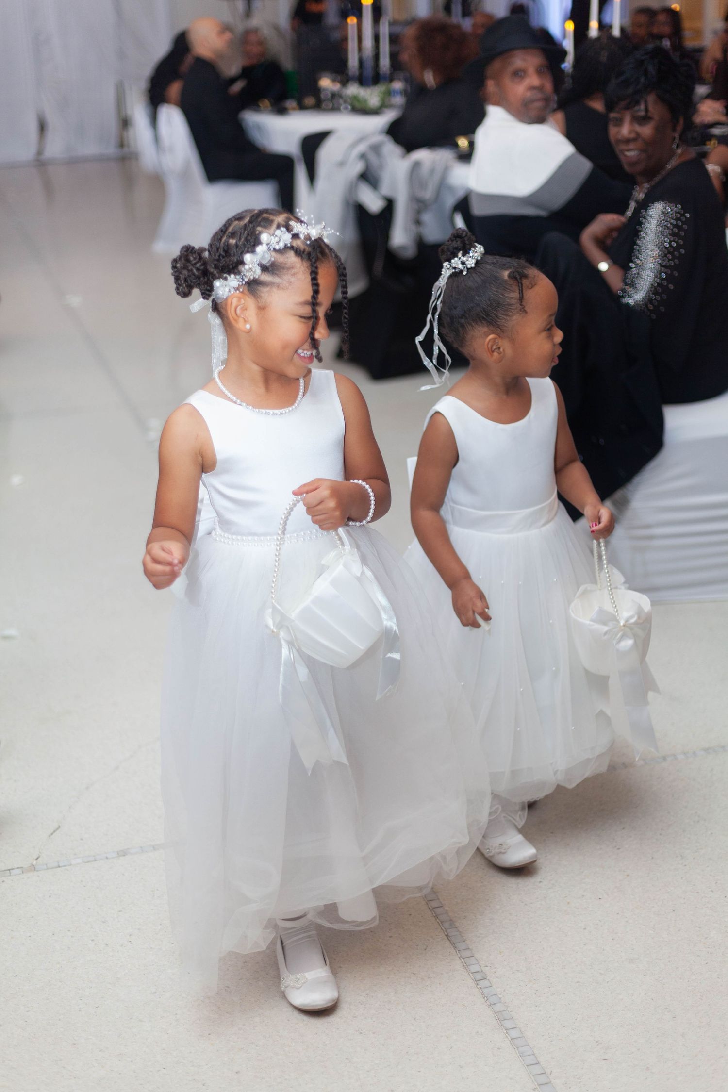 Two young flower girls in white dresses and headpieces walk together at a formal event.