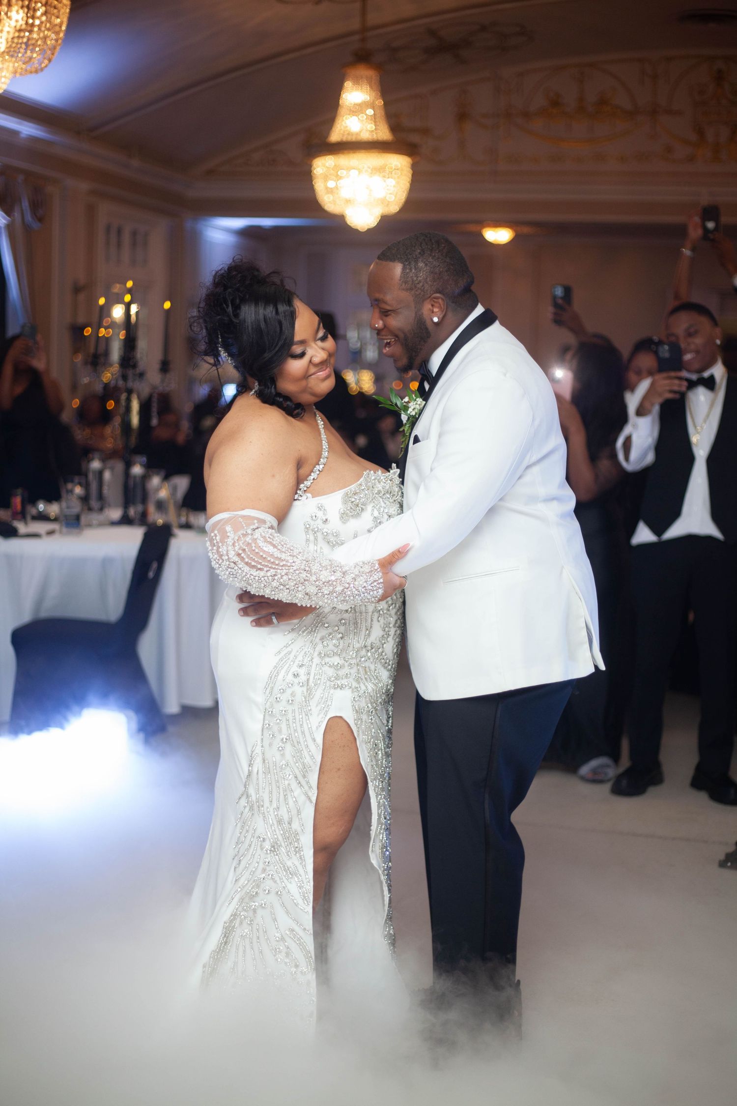 Couple shares an elegant dance in a grand ballroom with crystal chandeliers and white decor.