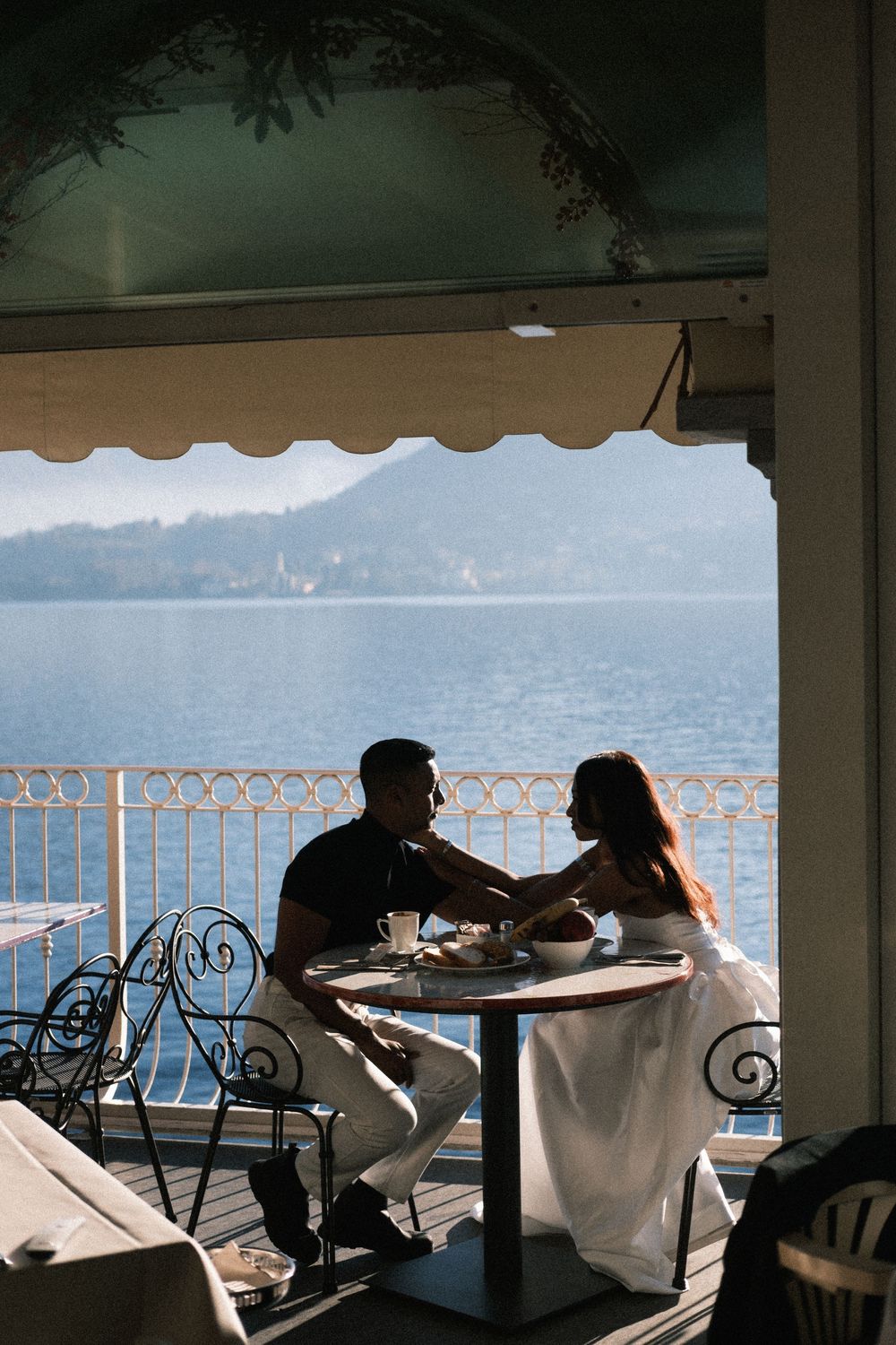Silhouetted diners enjoy a meal at an elegant waterfront restaurant with ornate railings overlooking a scenic lake view.