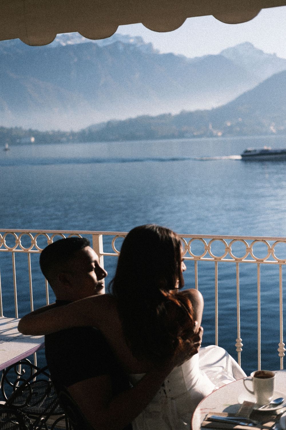 Two people relax at a lakeside cafe terrace with mountains and a boat visible across the tranquil water.