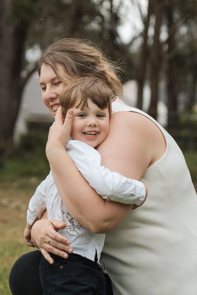 A parent in white embraces and plays with a laughing child in a white shirt outdoors among trees during a family photoshoot in Gables Box hi