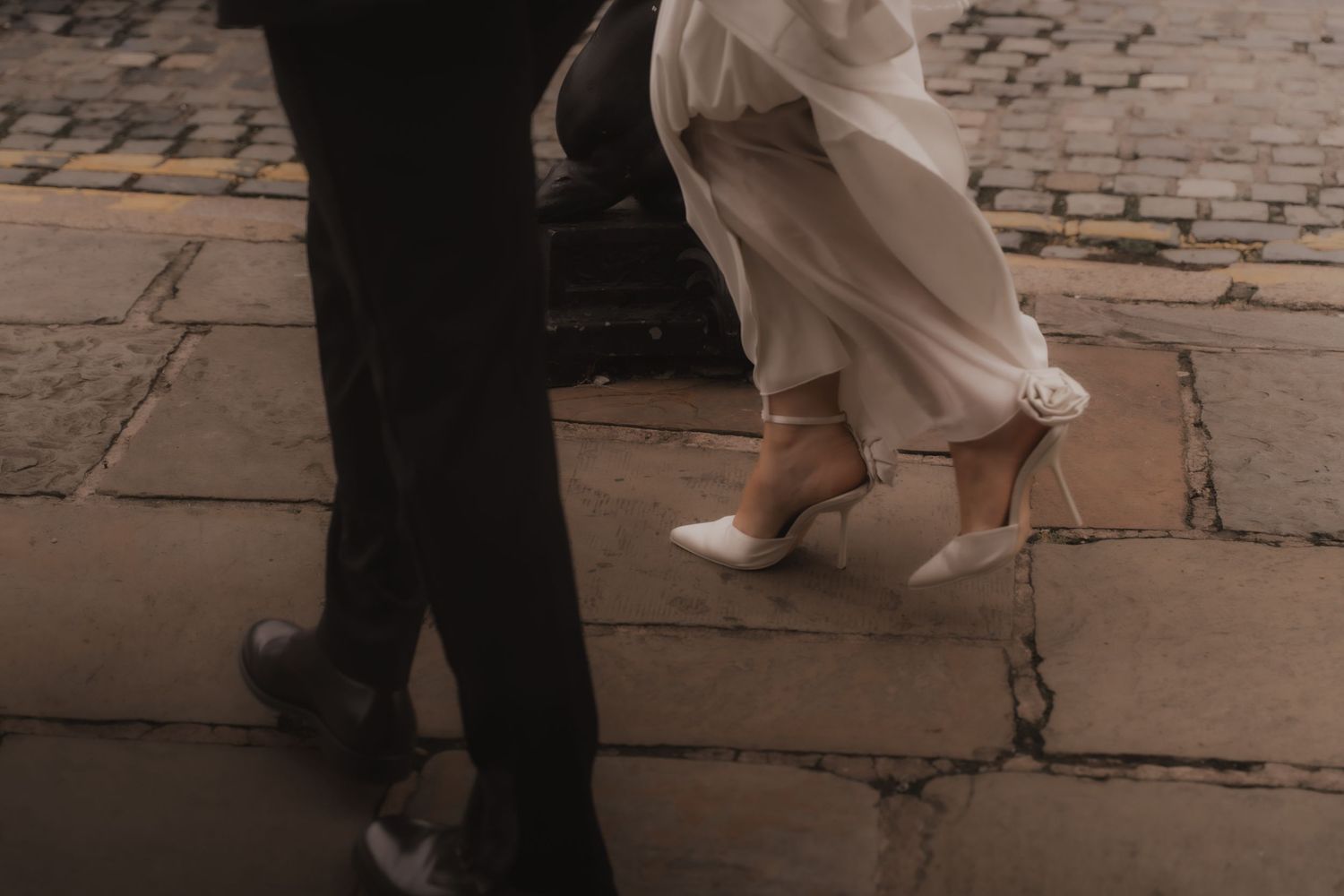 Close-up of feet walking on cobblestone pavement showing black shoes and white dress hem in motion.
