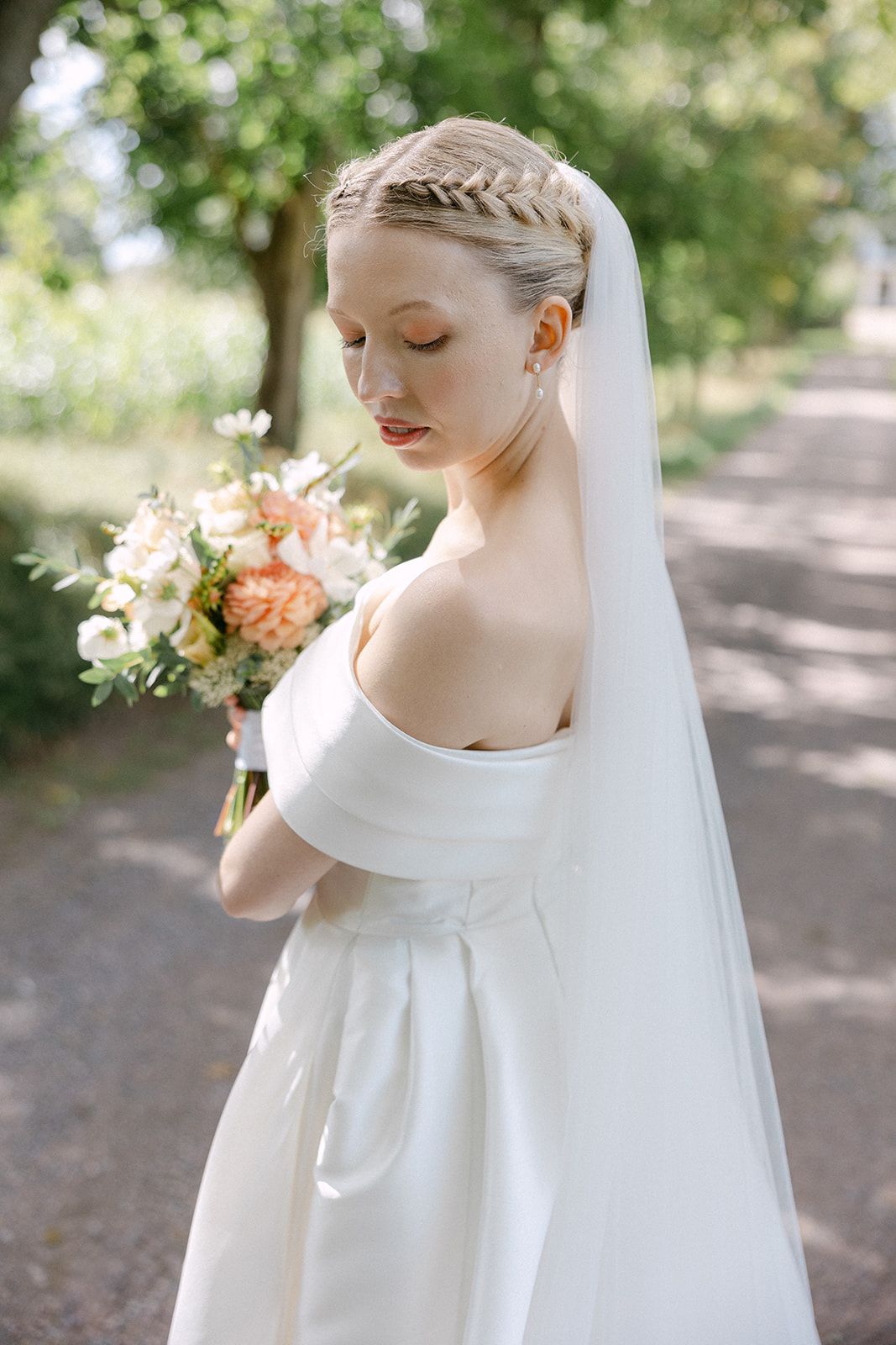 A bride in an elegant white wedding dress with off-shoulder sleeves poses gracefully in a garden setting.
