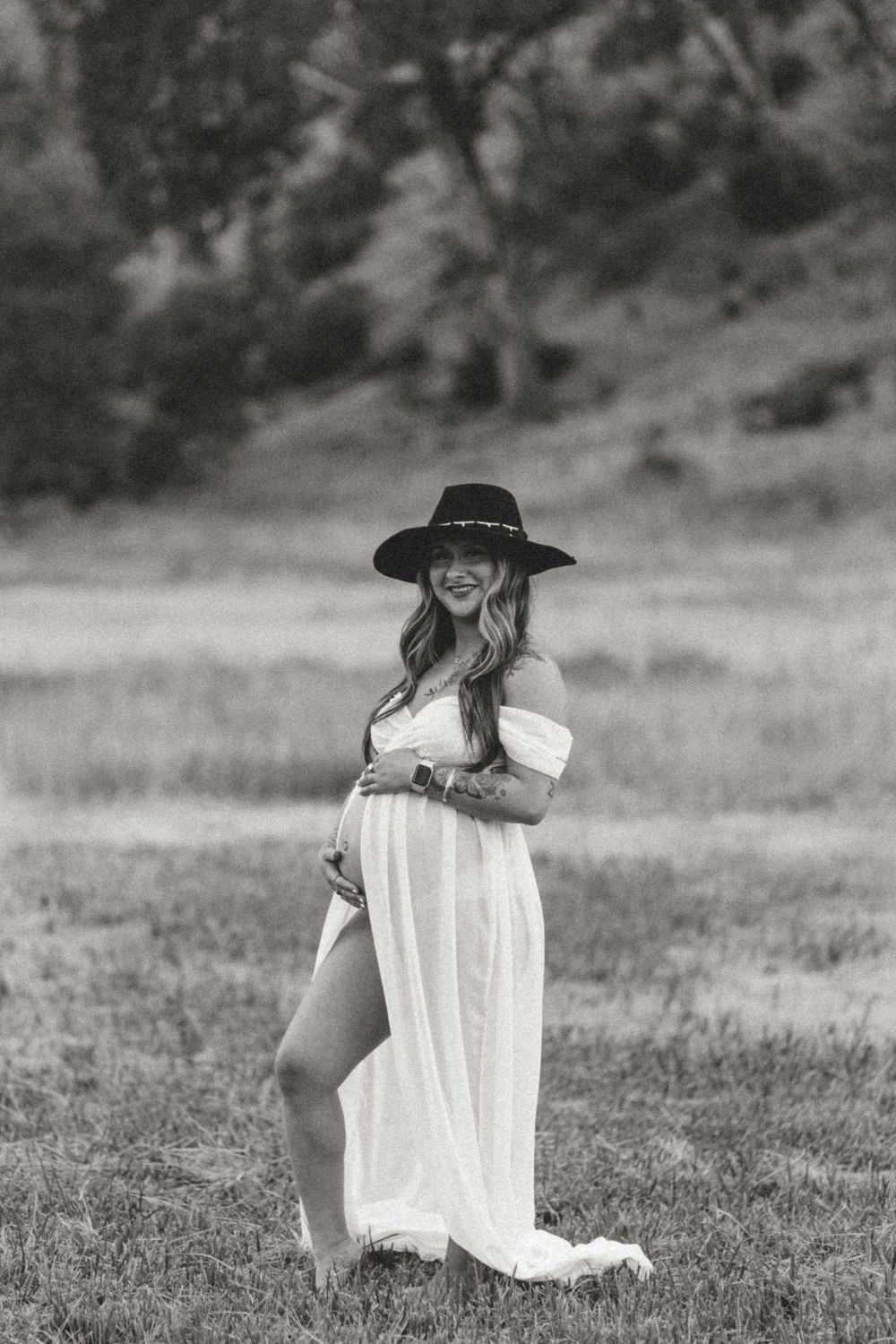 Black and white maternity photo of a woman in a flowing white dress and black hat standing in a grassy field.