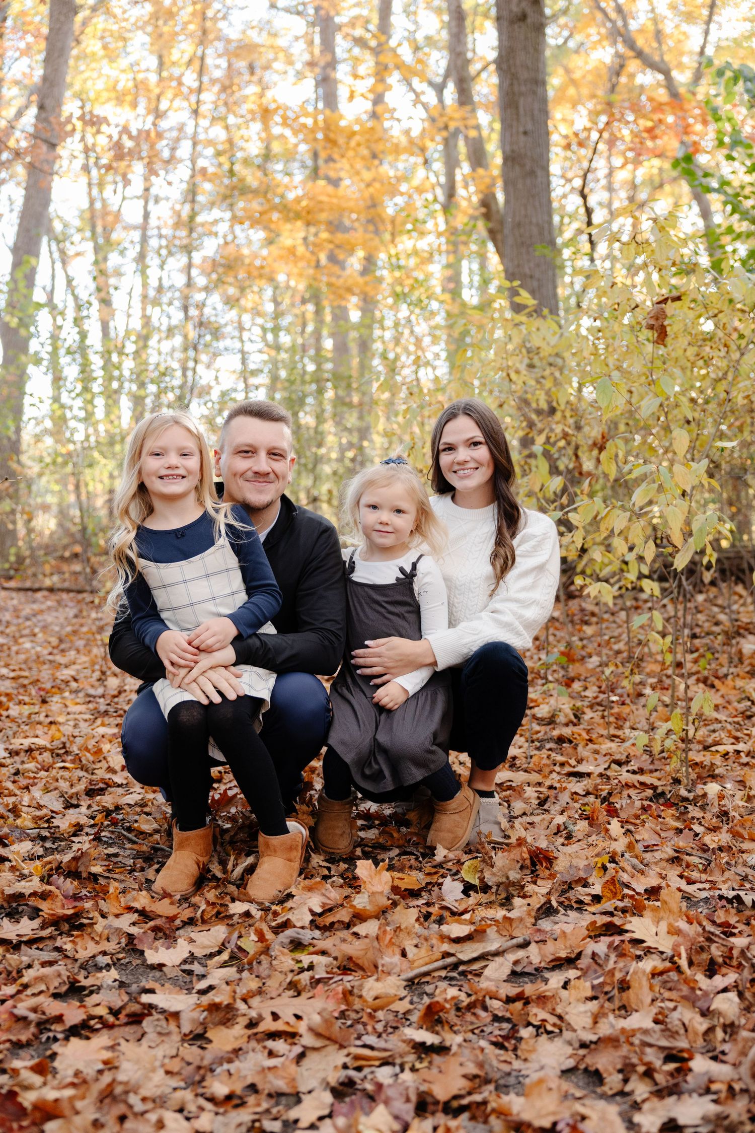 A family sits together on autumn leaves in the woods during a fall photo session.