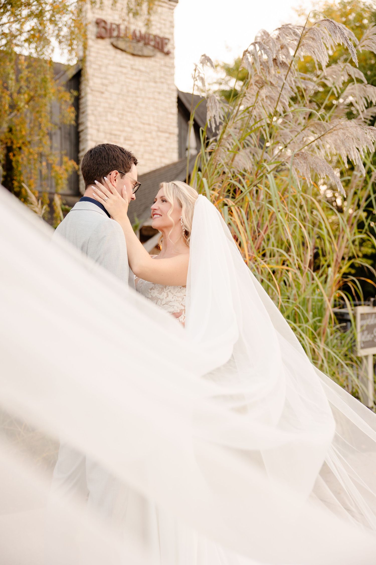 A romantic wedding couple embraces outdoors with a flowing white veil creating an ethereal effect in autumn light.