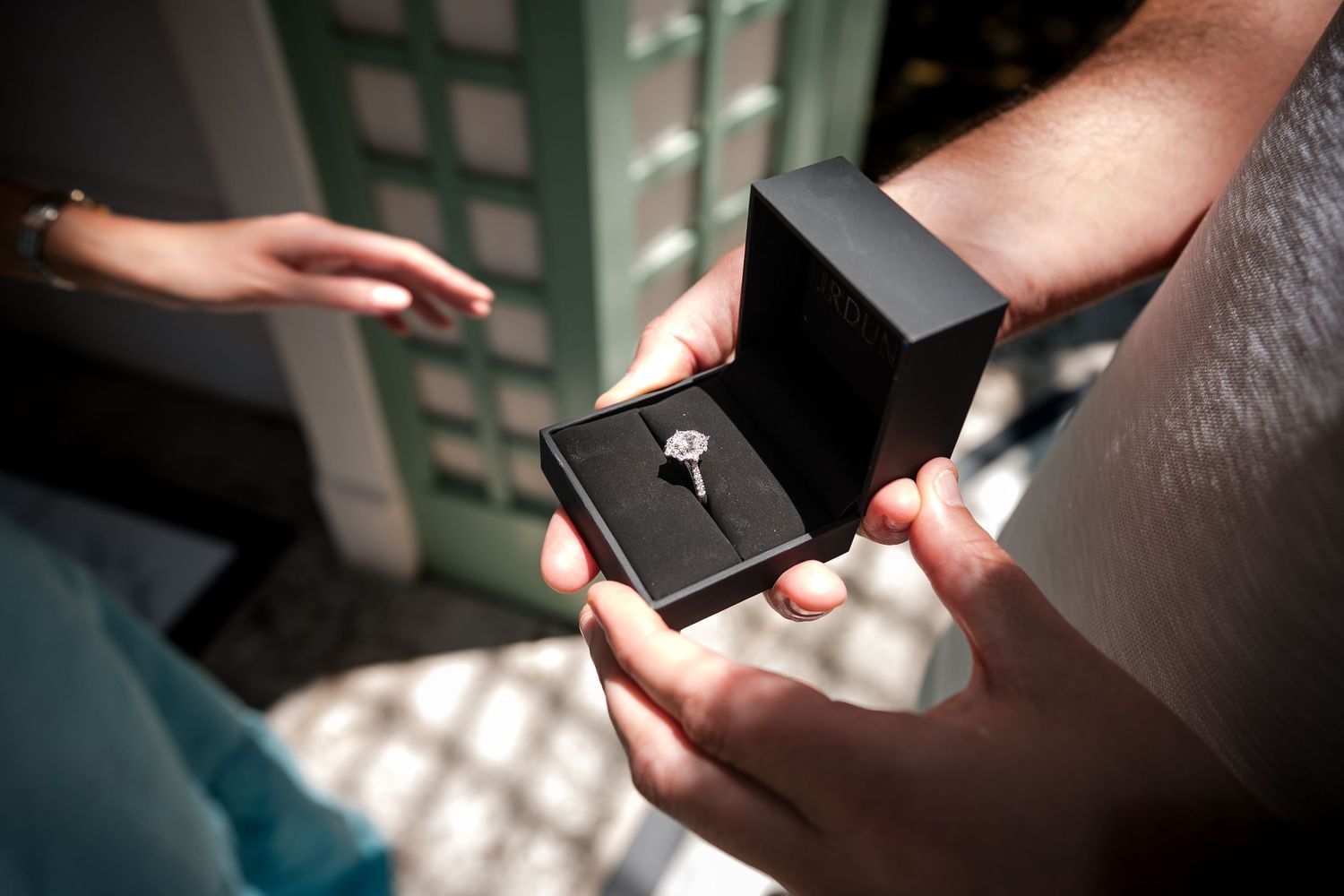 Person holding open black jewelry box with diamond engagement ring while proposing to partner outdoors.