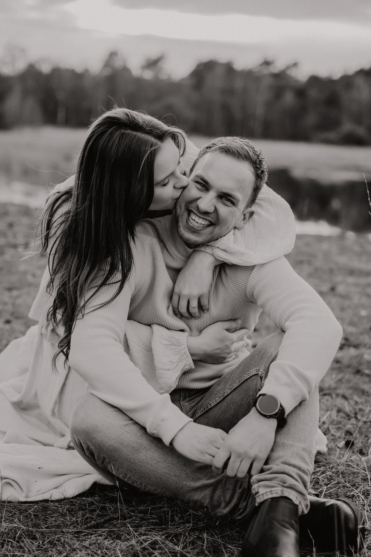 A couple laughs and embraces while laying in a grassy field in black and white photography.