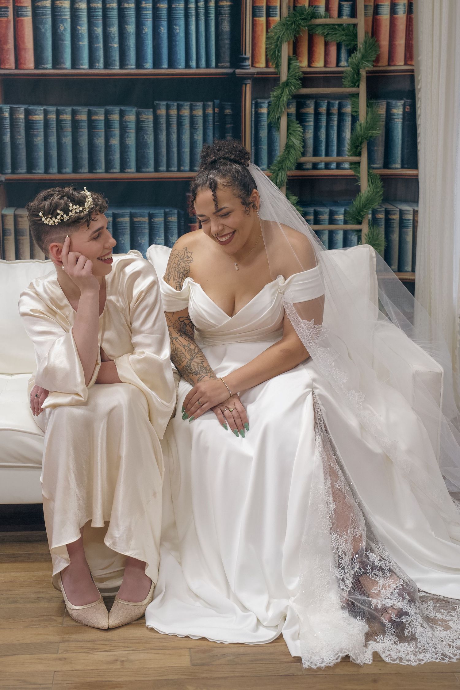 Two brides in white wedding dresses sit close together on a wooden floor in front of a wall of books.