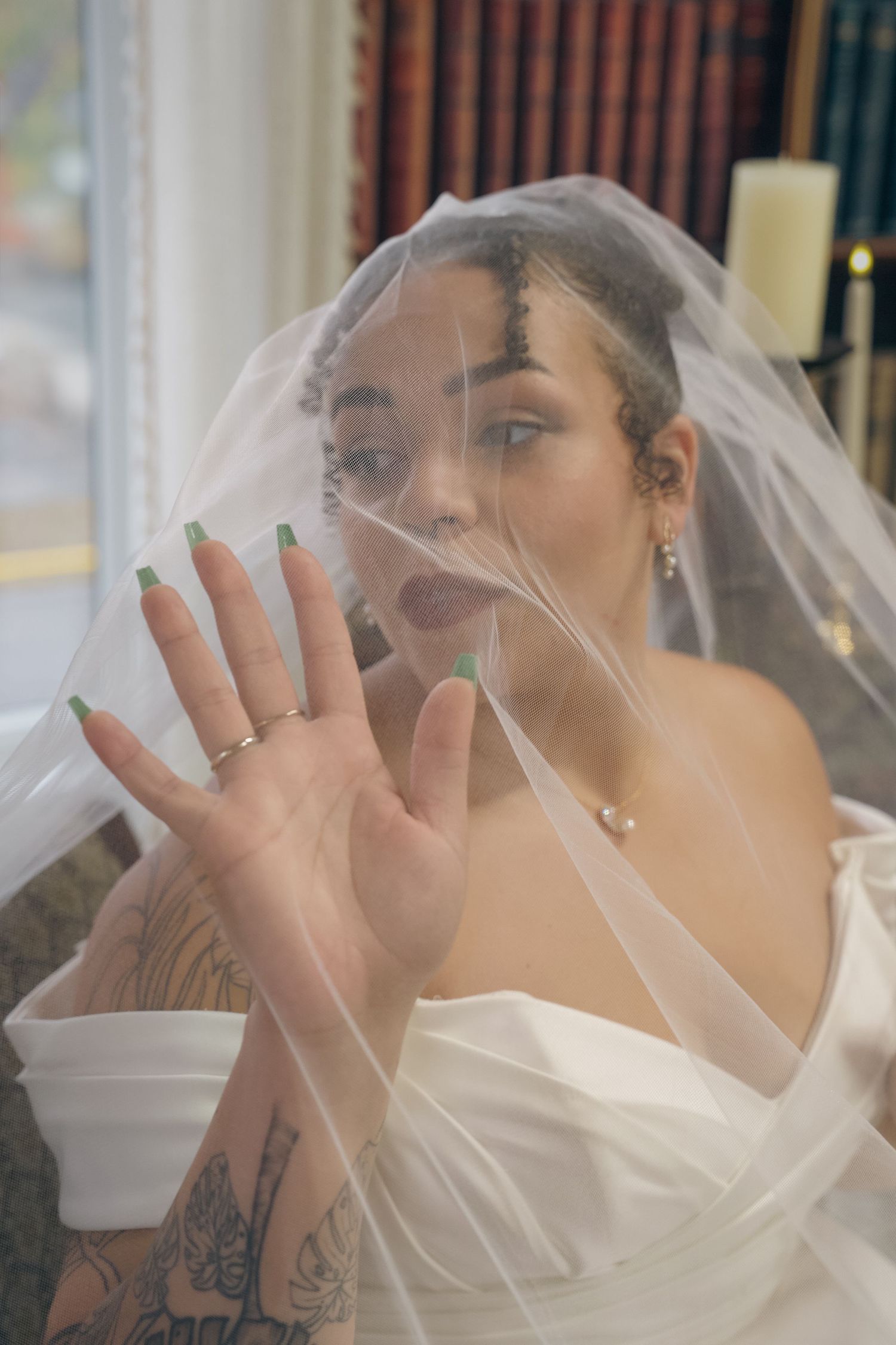 A bride in a white wedding dress and veil poses with her hand up against sheer tulle fabric, showing off her tattoos.