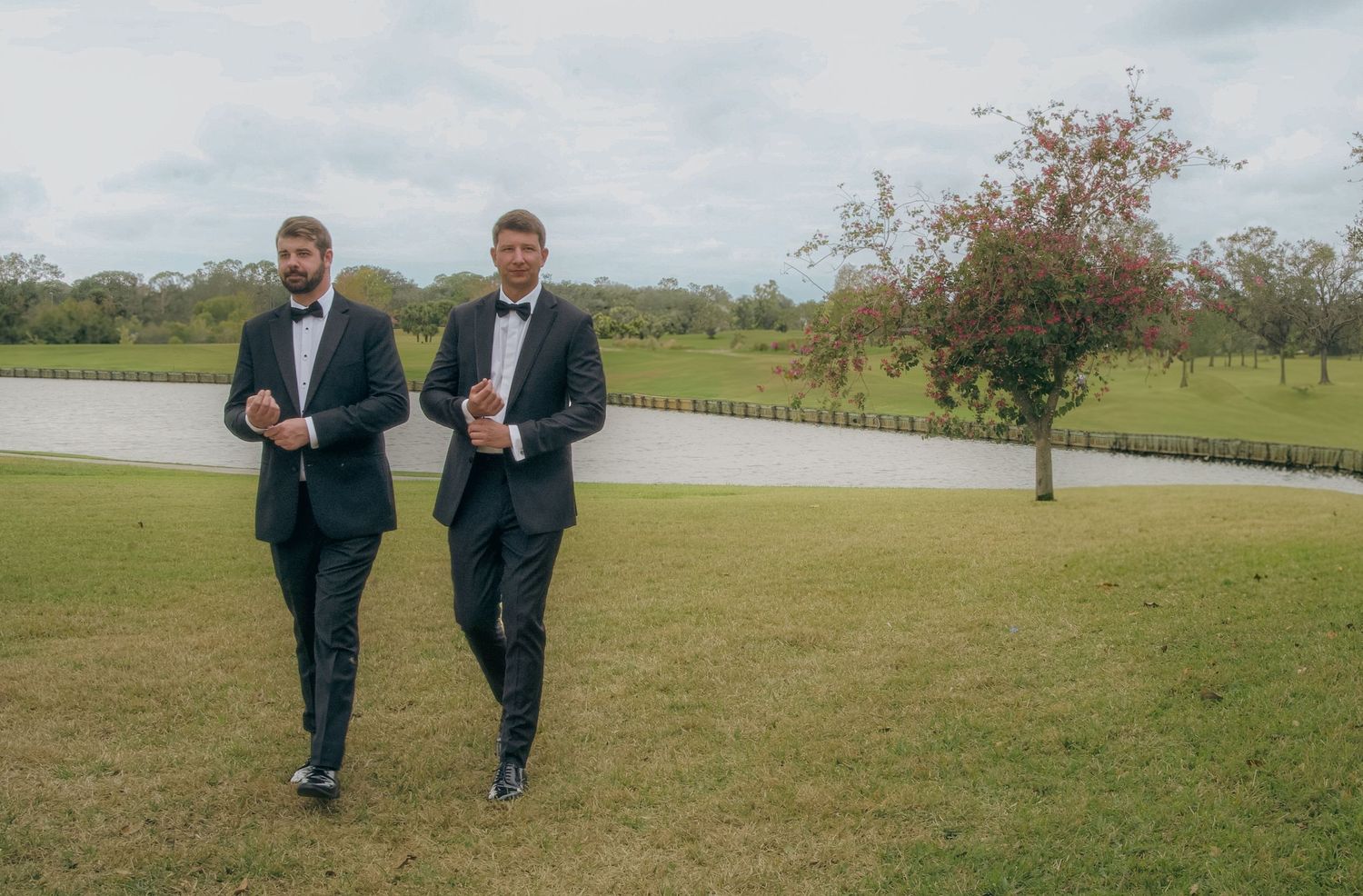 Two men in matching navy suits walk together across a green lawn on an overcast day.