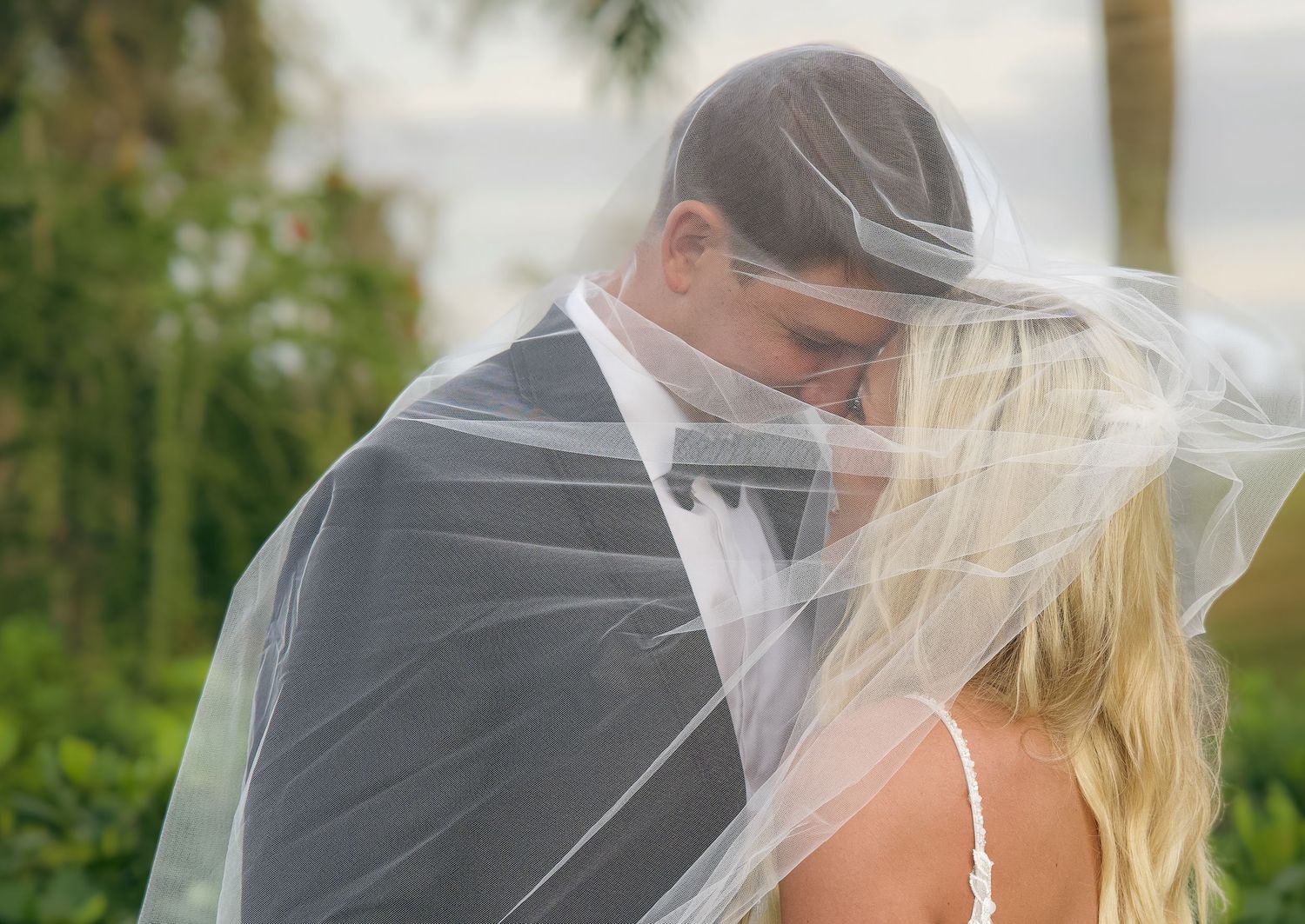 Bride and groom sharing intimate moment under flowing wedding veil outdoors with tropical greenery background.