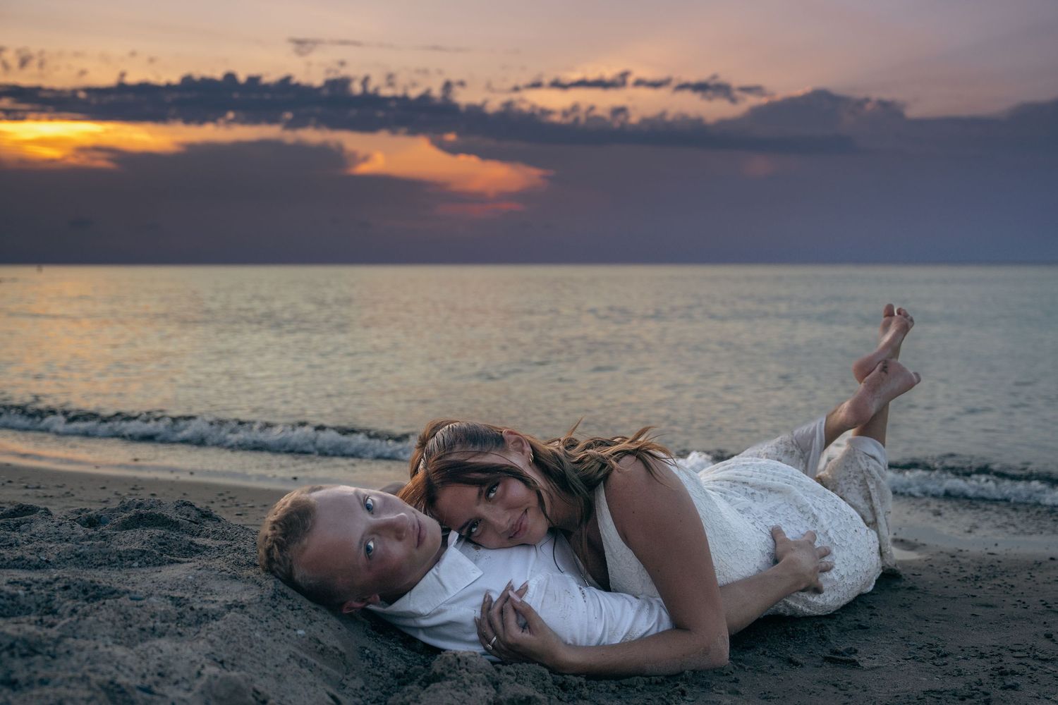 A couple lies together on a sandy beach at sunset with dramatic clouds over the ocean horizon.