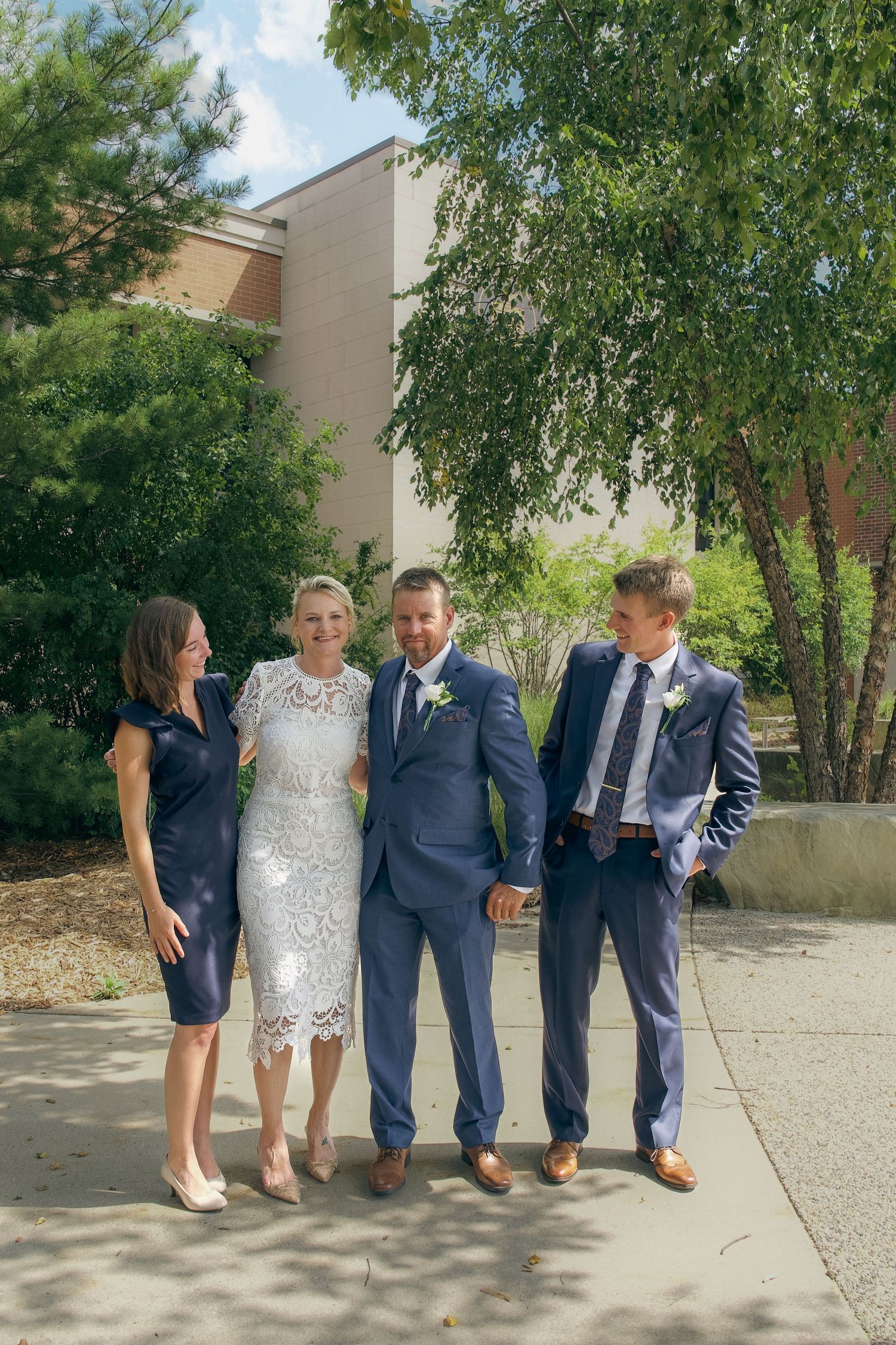 A wedding party poses outdoors wearing navy suits and formal attire while sitting on a low concrete wall.