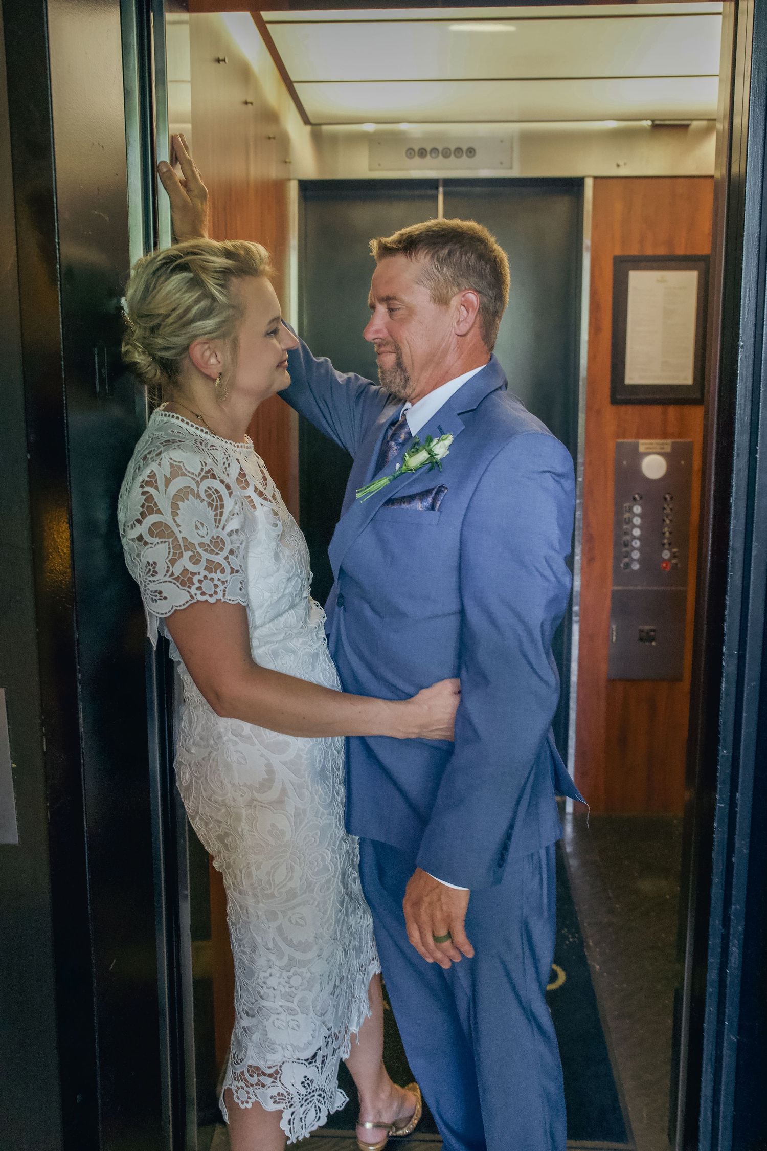 A couple shares an intimate moment in an elevator, with the bride wearing a lace dress and groom in a blue suit.