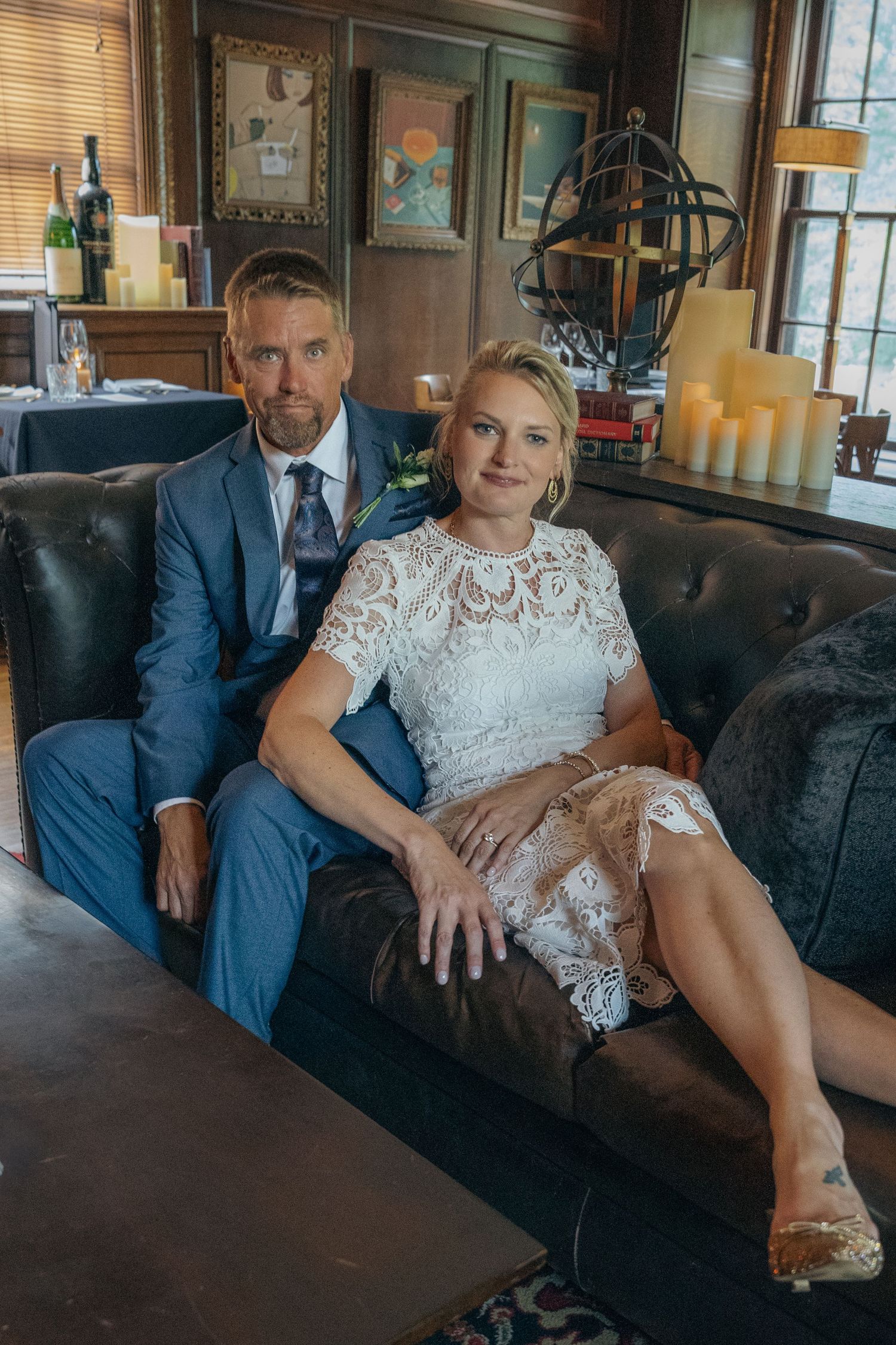 Couple in wedding attire poses together on a vintage leather couch in an elegantly decorated indoor setting.