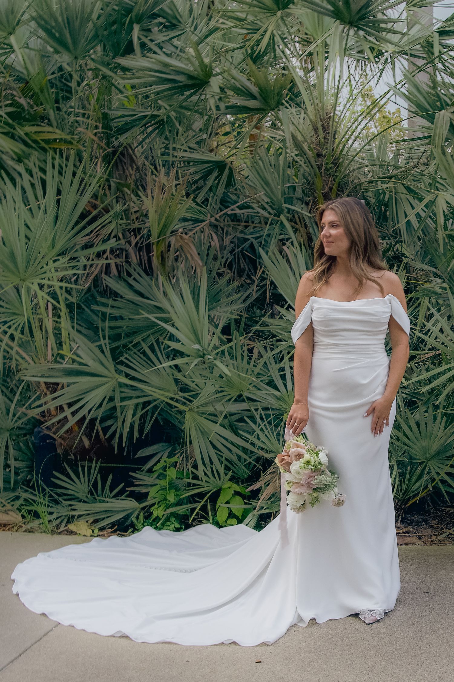 A flowing white wedding dress with a long train is showcased against a backdrop of tropical palm plants.