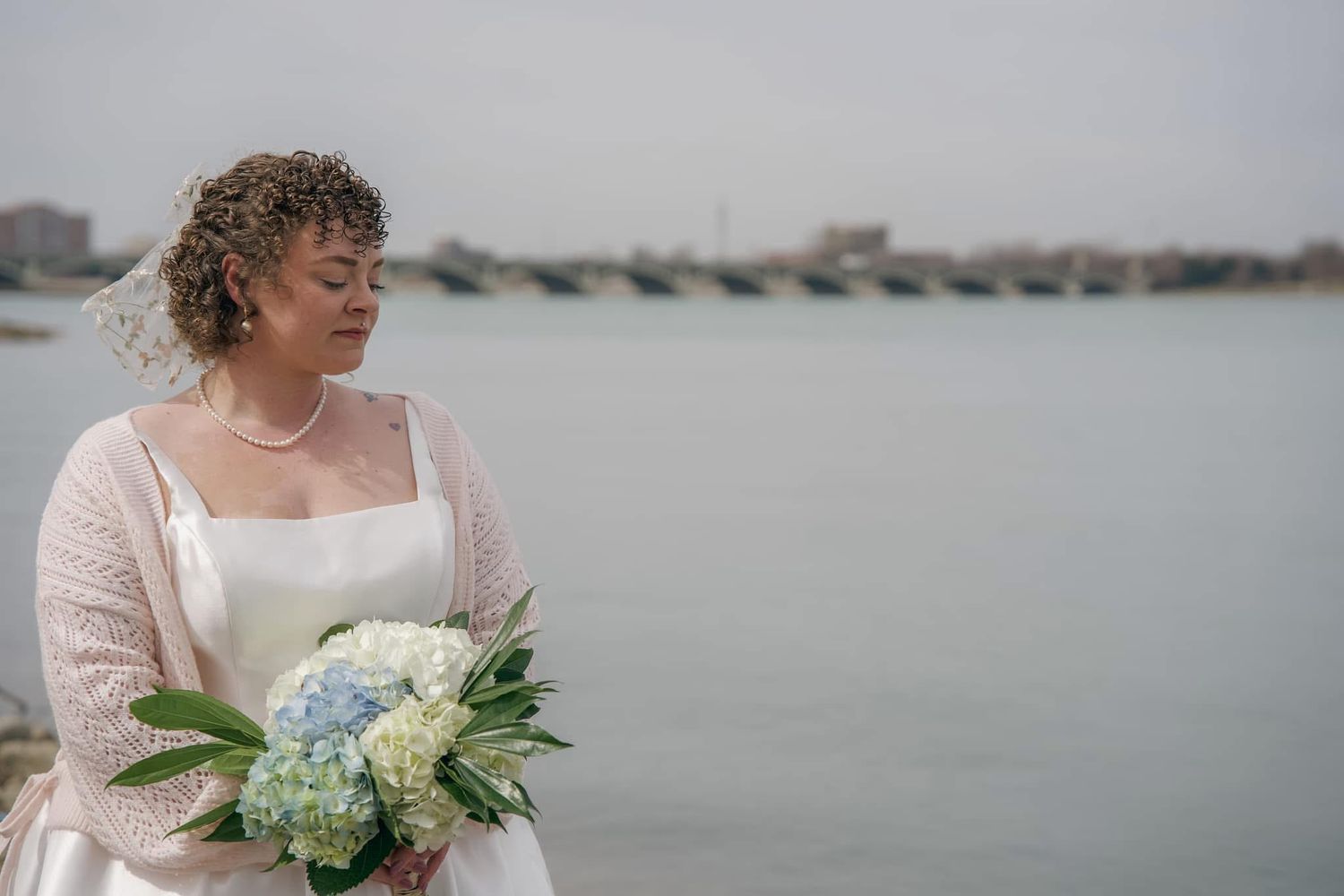 Elegant bride in lace sleeves holds a soft blue and white hydrangea bouquet against a coastal backdrop.