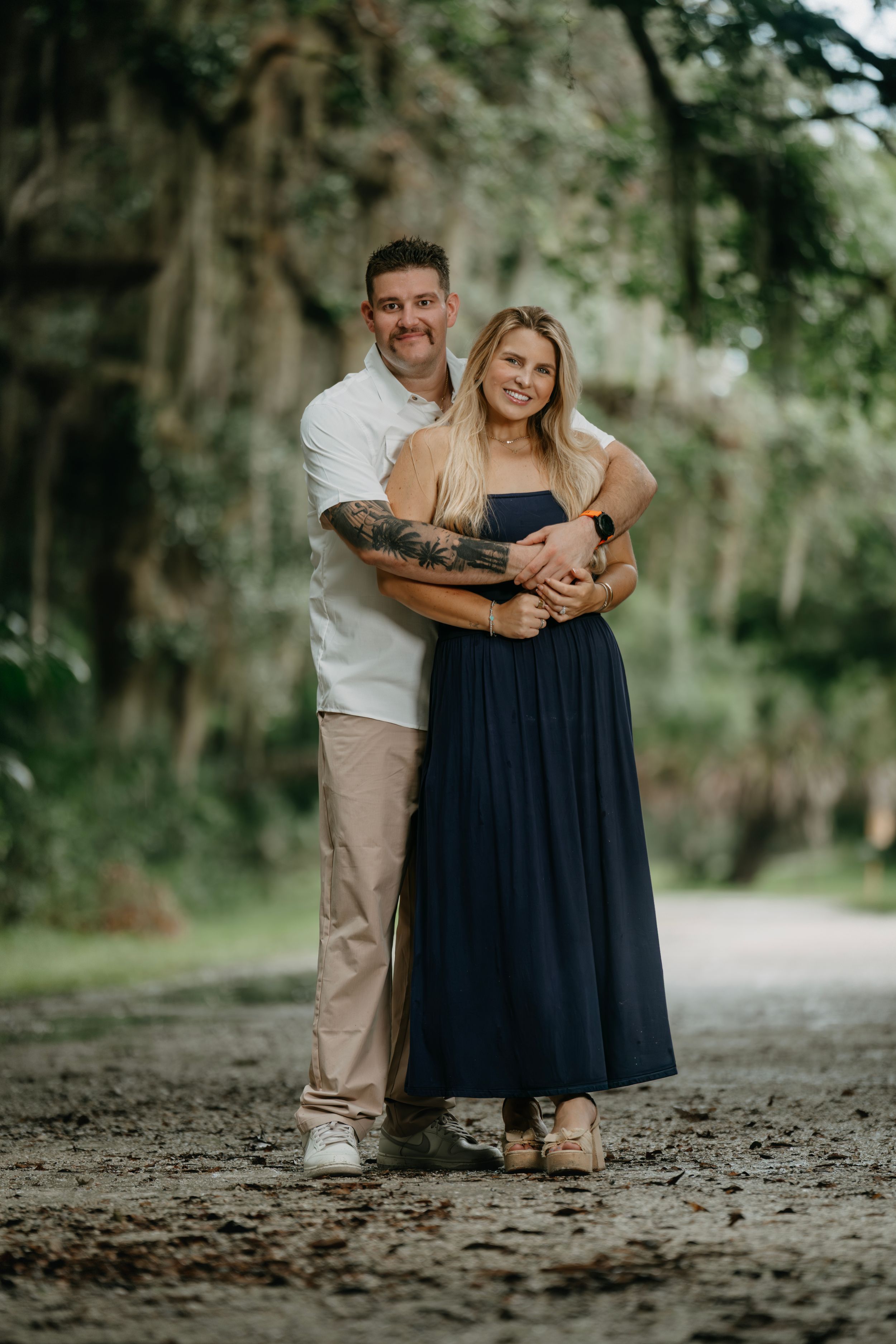 Couple embracing on a scenic path surrounded by lush green trees, woman in navy maxi dress, man in white shirt.