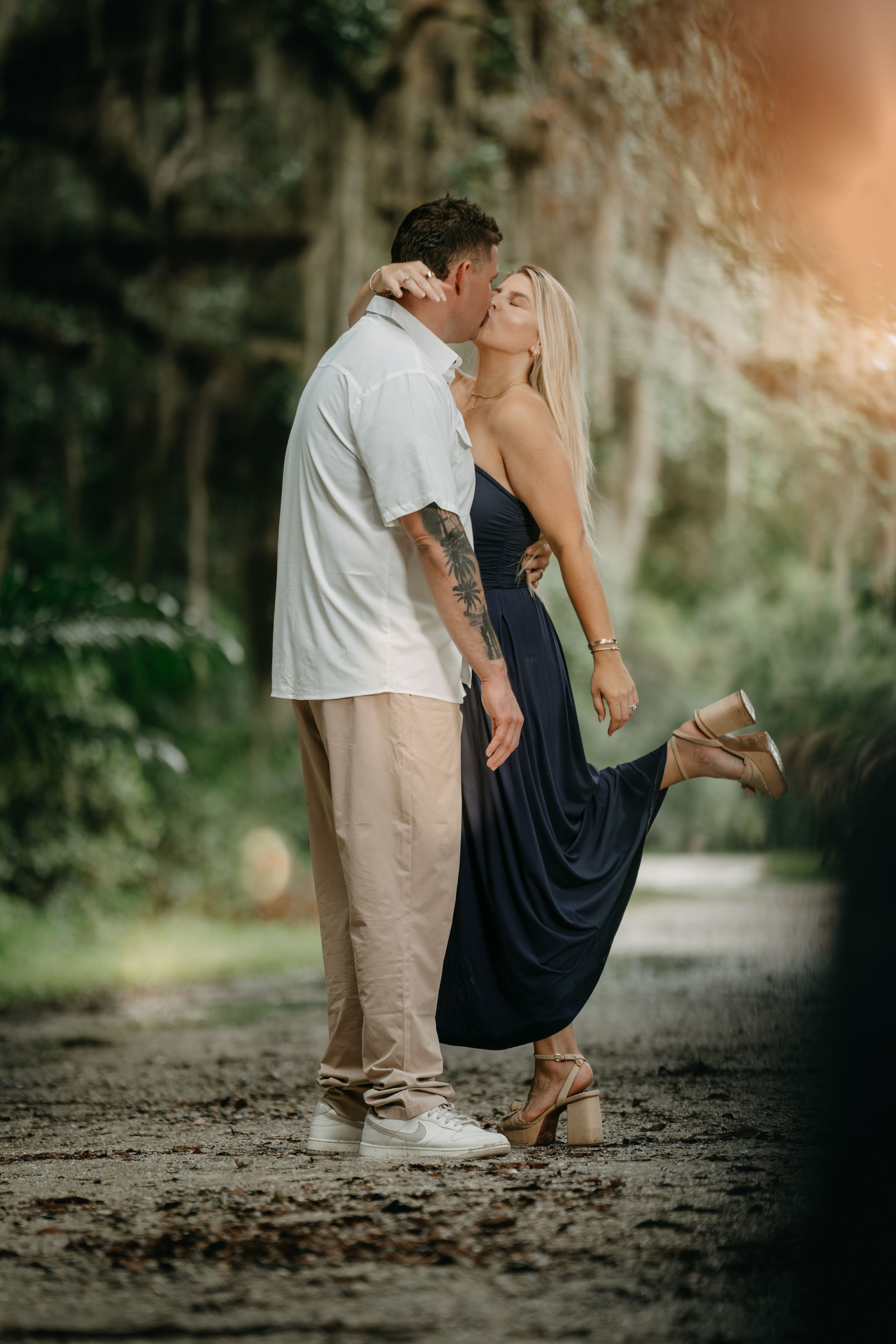 A couple shares a romantic kiss on a scenic forest path, the woman in a navy dress lifting one heel.