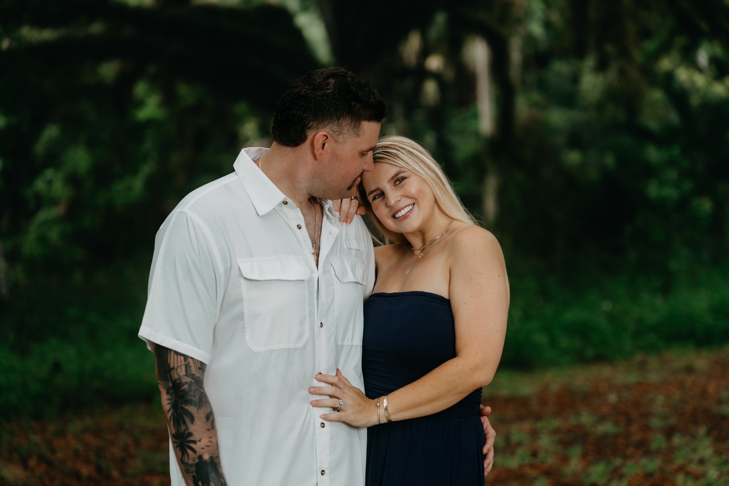 A tattooed man in a white shirt kisses a smiling blonde woman in a navy dress outdoors.