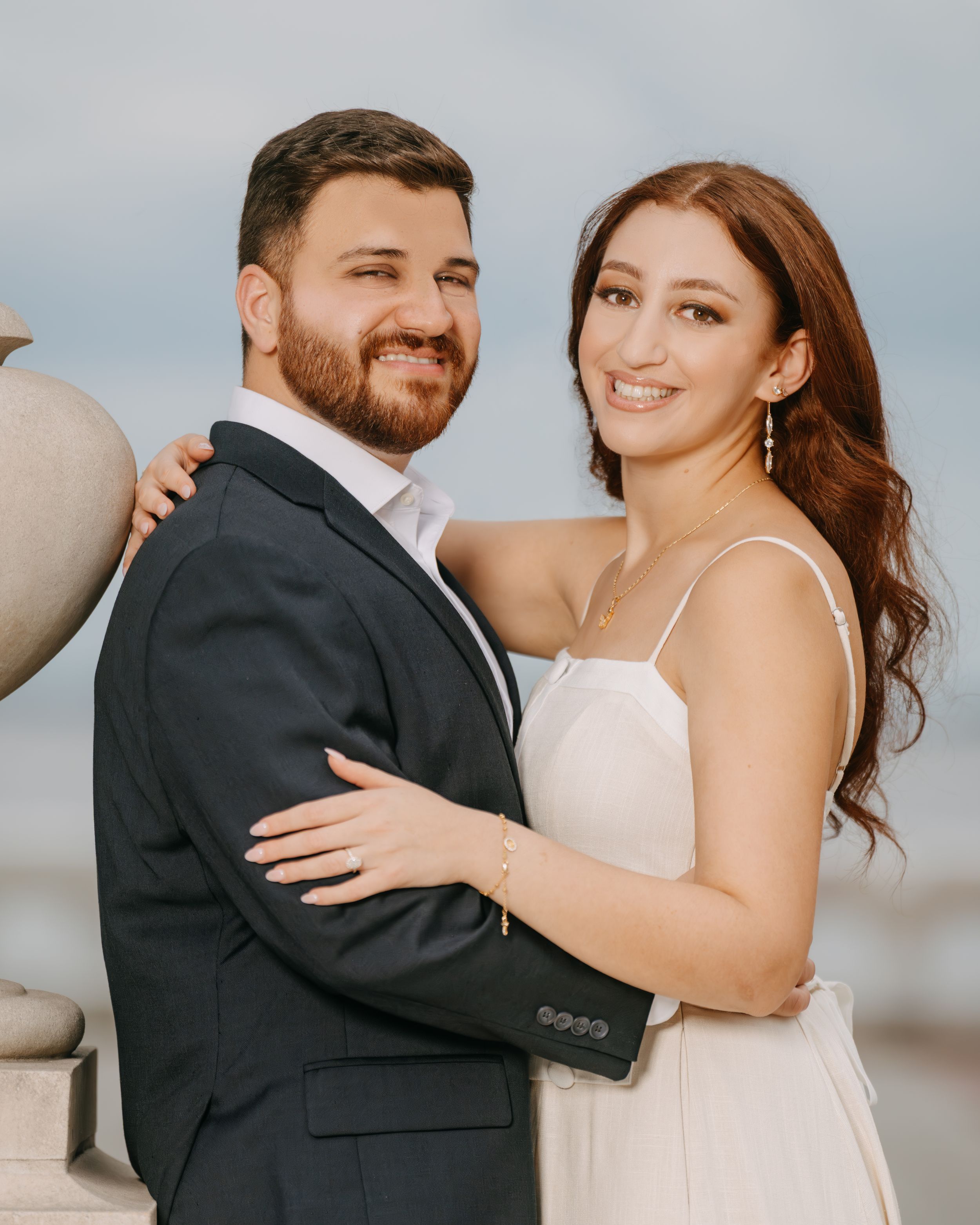 A smiling couple in formal attire poses closely together outdoors, the man in a dark suit and woman in a white dress.